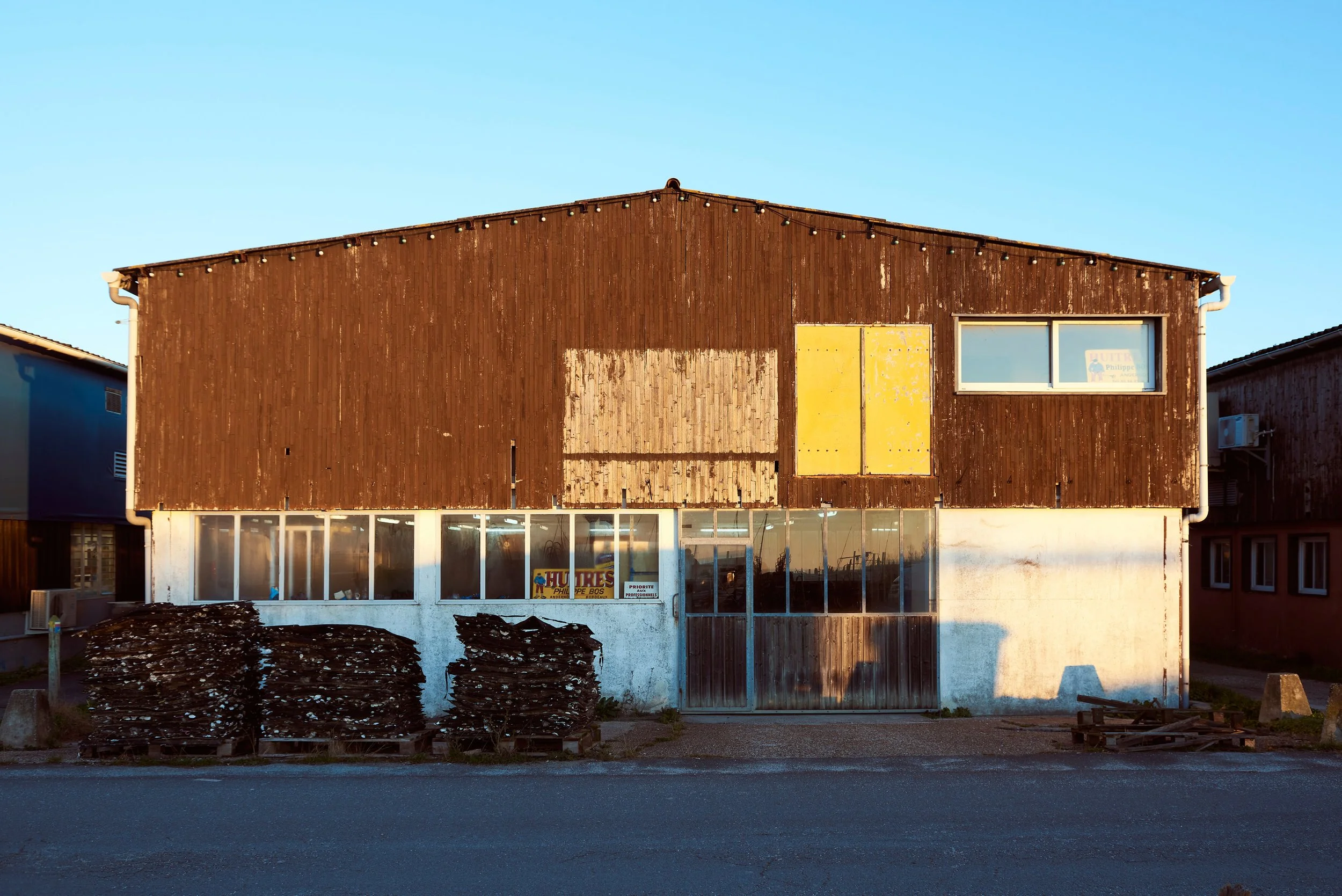 Bâtiment en bois avec des fenêtres et une porte, devant une zone de débris et de piles de bois, sous un ciel clair.
