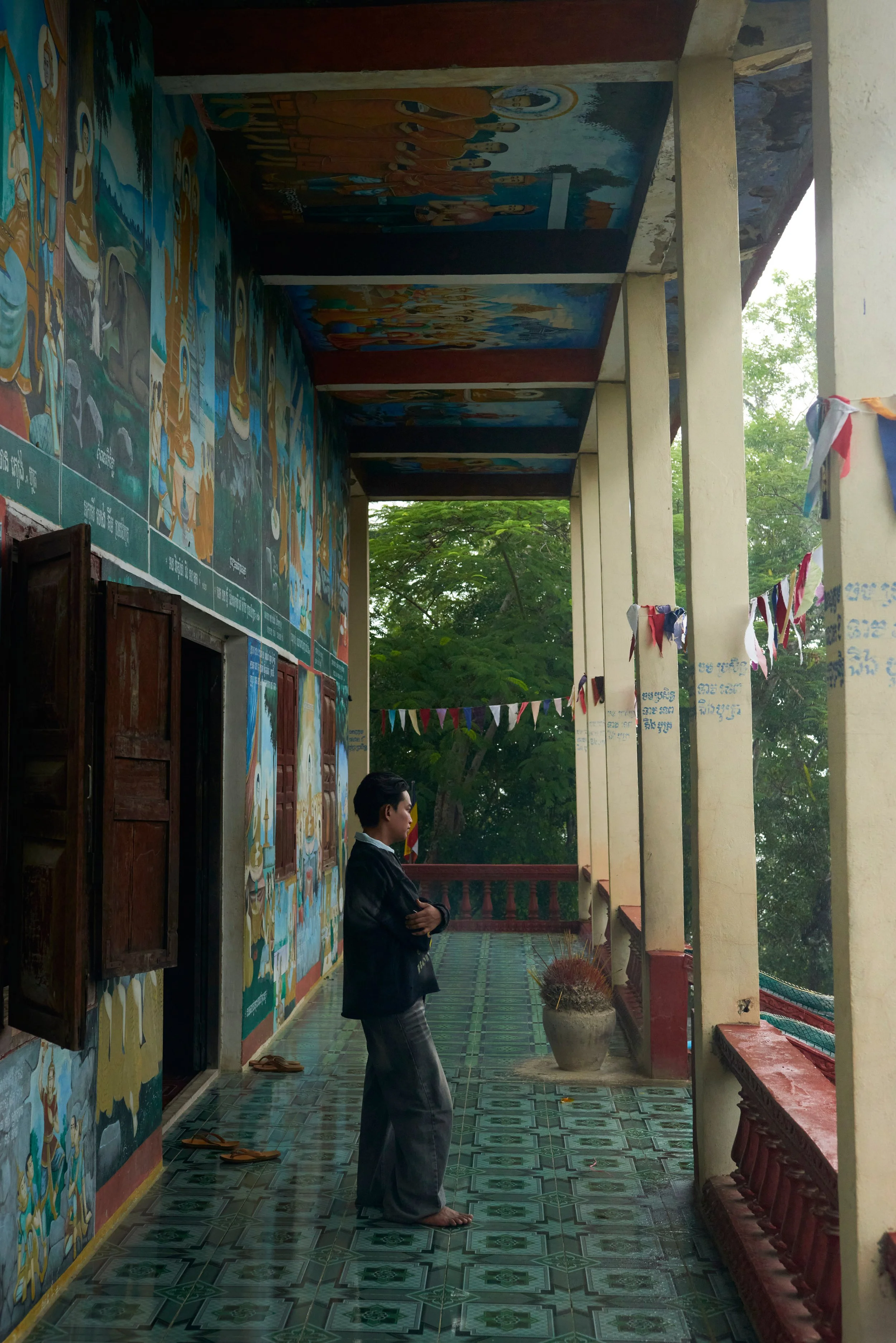 Une personne en train de prier dans un espace intérieur d'une église ou d'un temple. La personne est debout, pieds nus, avec les bras croisés et regardant vers l'avant. Le mur derrière elle est décoré de peintures religieuses colorées, avec des colon