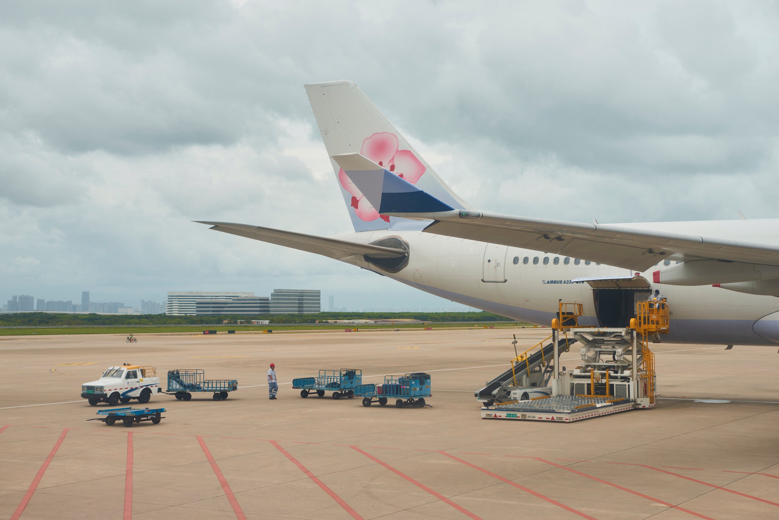 Avion de ligne à l'aéroport, avec un personnel au sol et des équipements pour le traitement des bagages, sous un ciel nuageux.
