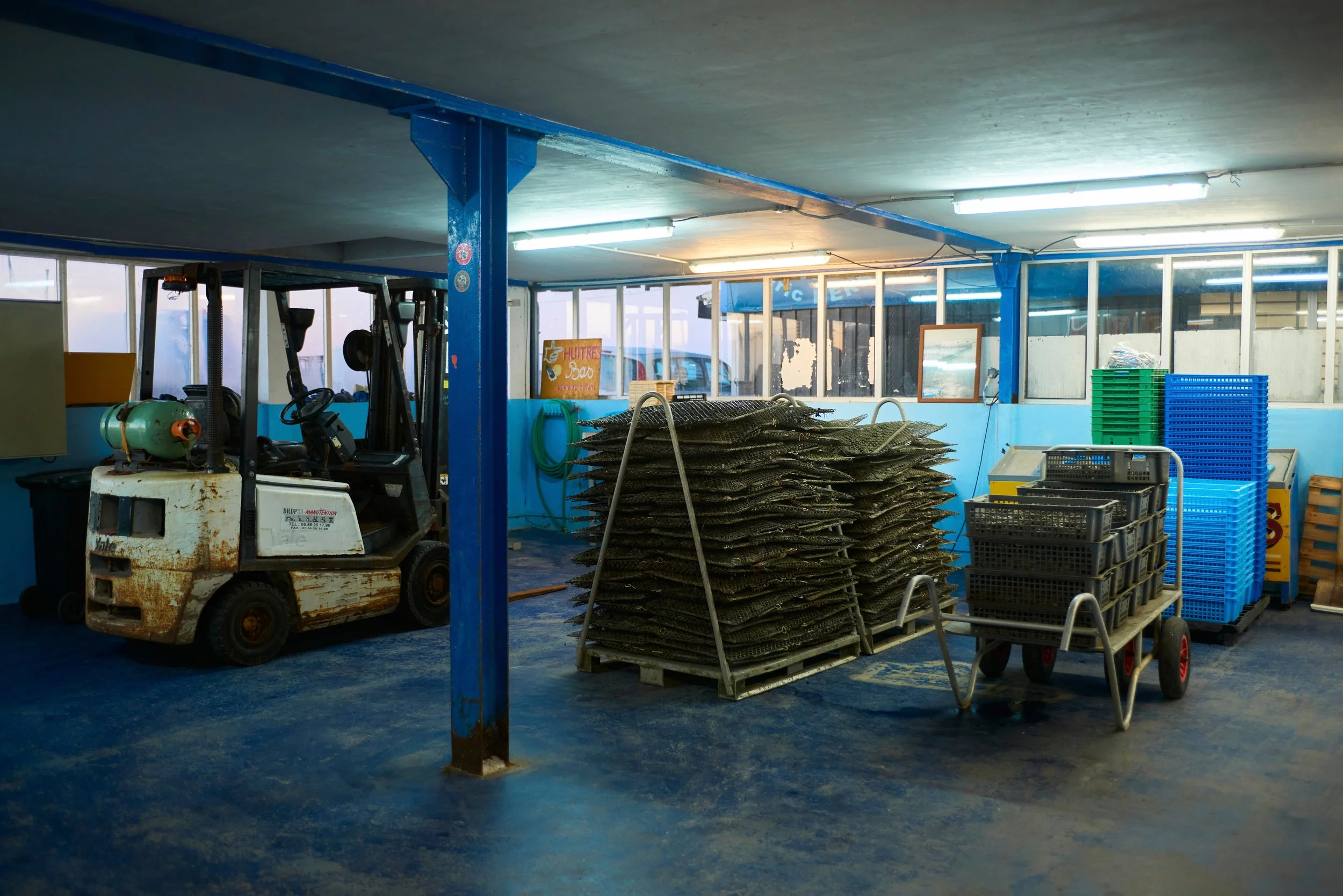 Intérieur d'un entrepôt avec une montagne de caisses en plastique, un chariot, une machine de manutention rouillée, et une caisse en bois. Fenêtres et lumières fluorescentes. Signes et éléments de stockage en arrière-plan.