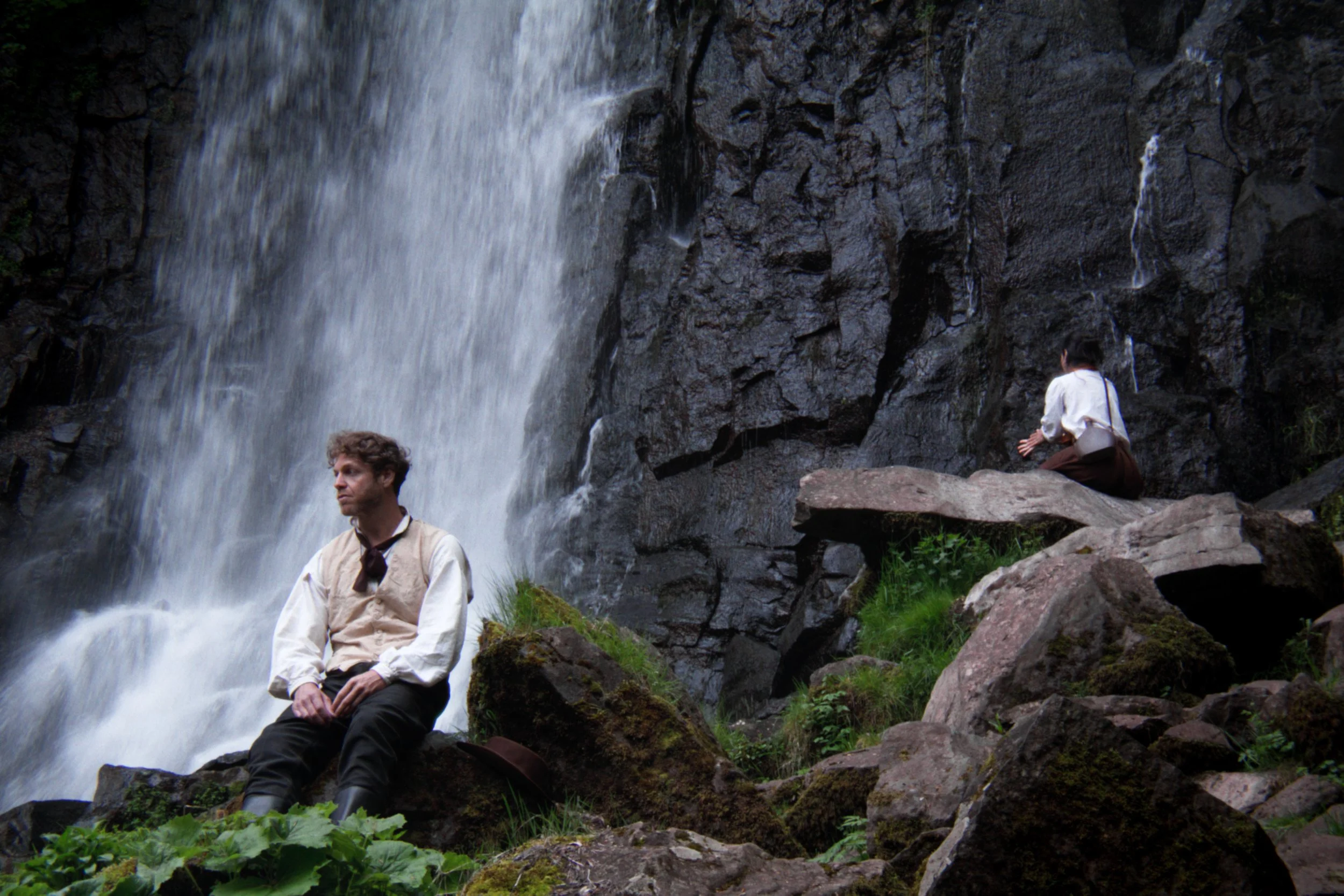 Deux personnes près d'une cascade rocheuse, une homme assis en avant et une femme en arrière, tous deux dans un environnement naturel avec des rochers, de l'eau et de la végétation.