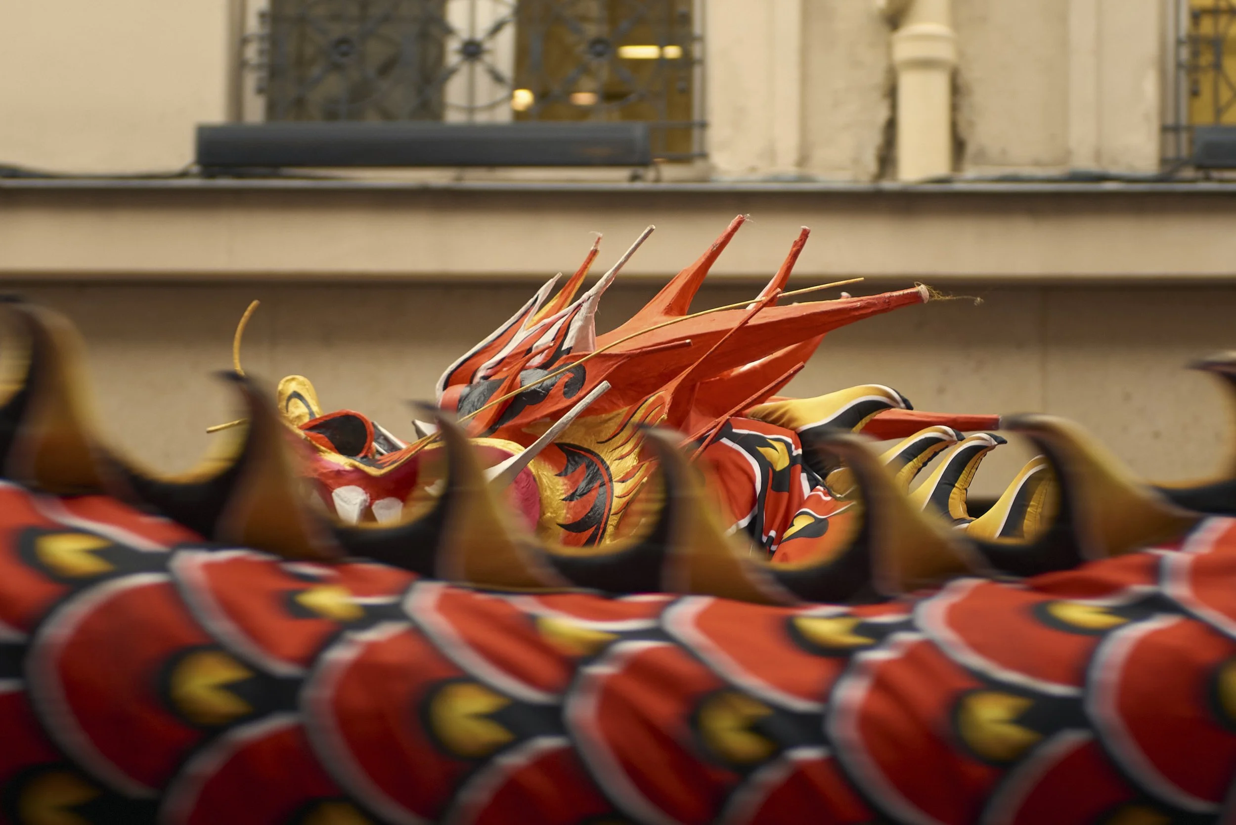 Une sculpture de dragon colorée exposée derrière une grille de protection. En arrière-plan, il y a une façade de bâtiment avec des fenêtres ornées. La photo est prise de près, mettant en évidence les détails de la sculpture.