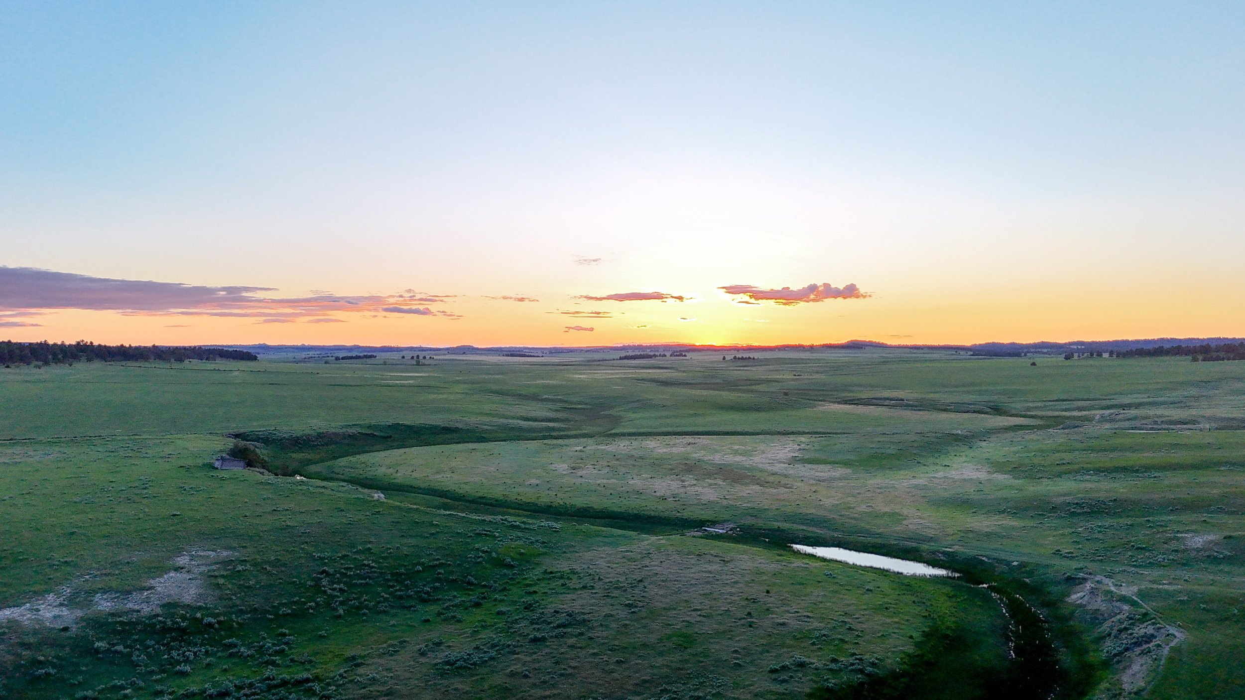 Scenic view of a vast green landscape with rolling hills and a small stream, under a clear sky with the sun setting on the horizon.