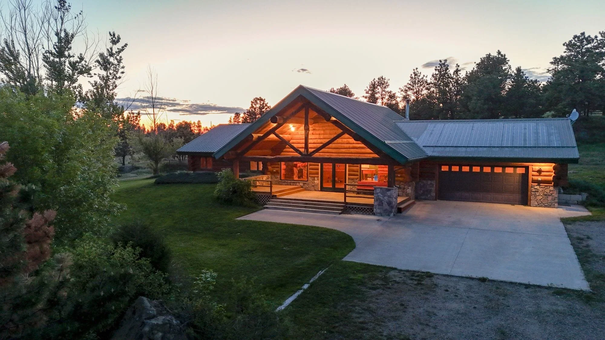 A log cabin-style house with a metal roof, porch lights on, and a driveway, surrounded by trees at sunset.