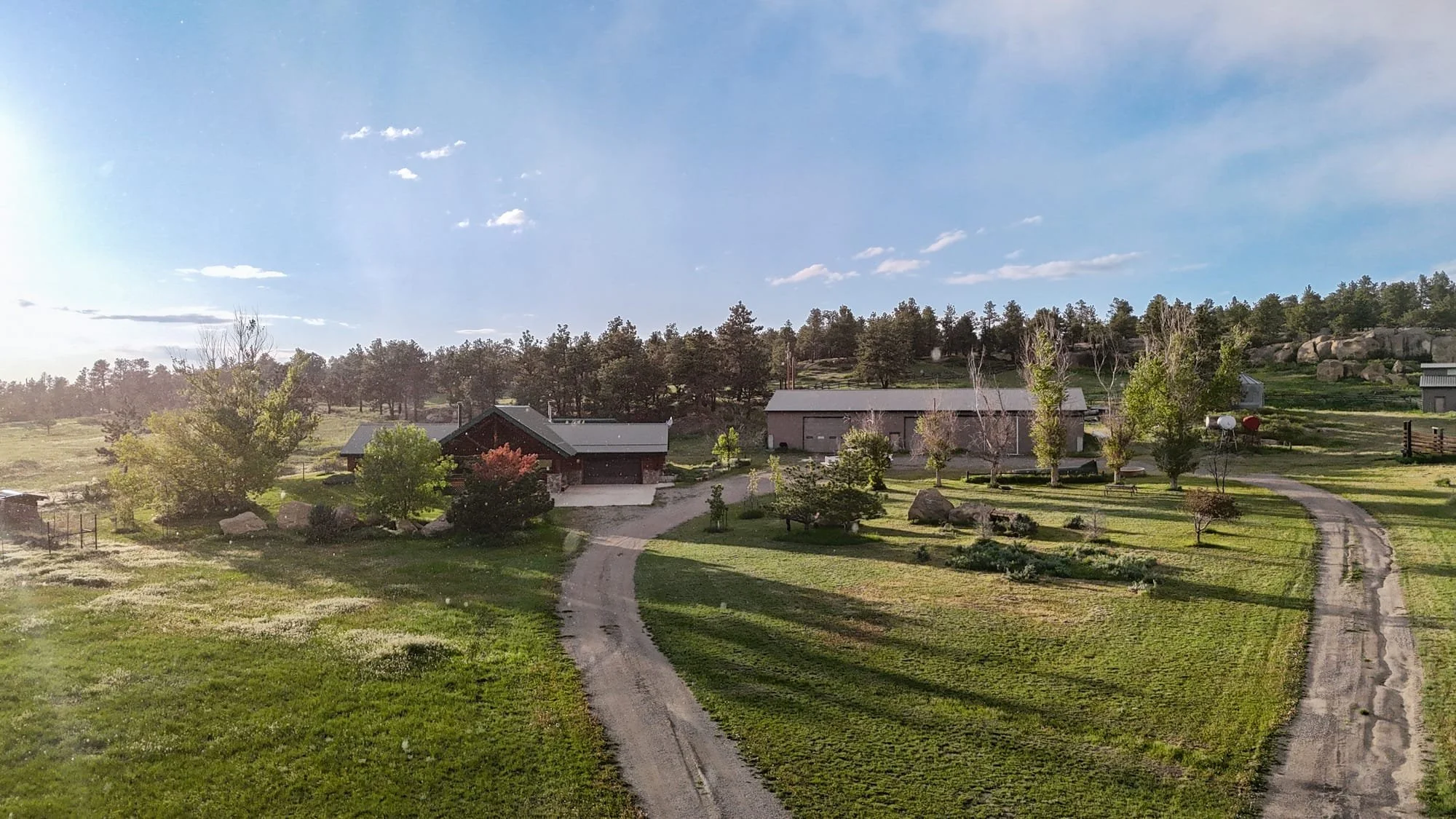 A rural landscape featuring a farmstead with a few buildings, surrounded by trees, green grass, and a winding dirt road on a sunny day with a blue sky and some clouds.