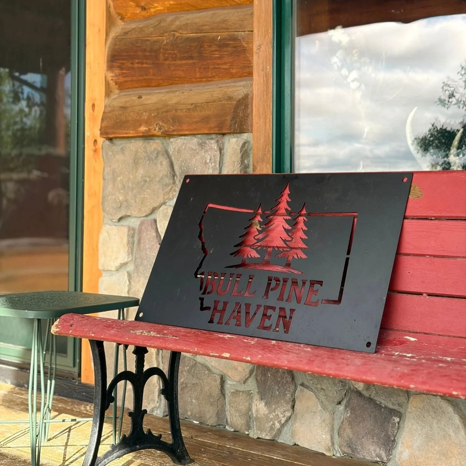 A black metal sign with cutout trees and the words 'Bull Pine Haven' leaning against a red wooden bench outside a building. The building has a stone and wood exterior with a large window reflecting the sky and trees.