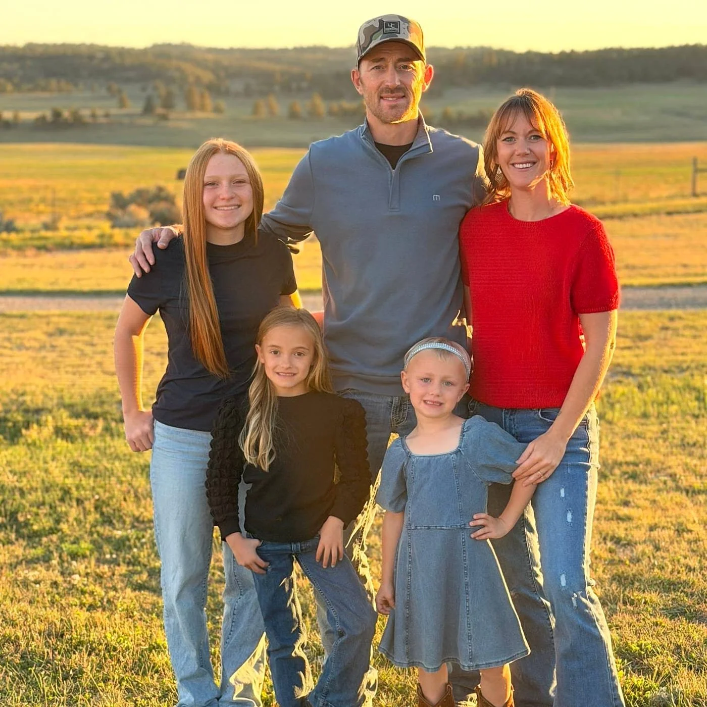 A family of six standing outdoors in a field during sunset, smiling at the camera. The family includes two adult women, one man, and three young girls. The background features rolling hills and trees.