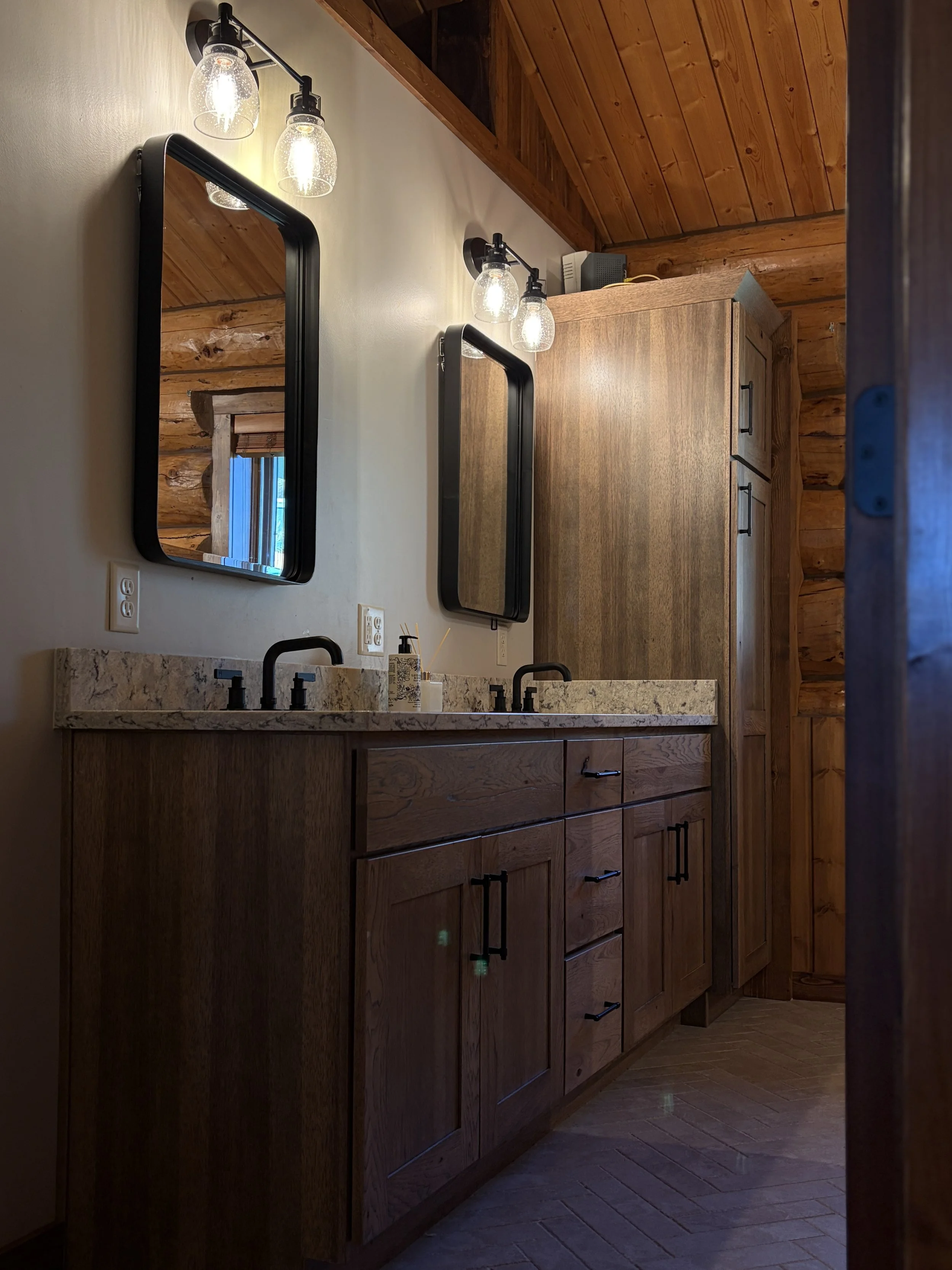 Wooden cabinet with black handles, marble countertop with a black faucet, and a mirror above. Two light fixtures with clear glass shades are mounted on the wall beside the mirror, illuminating the area. The ceiling is wooden, matching the rustic styl