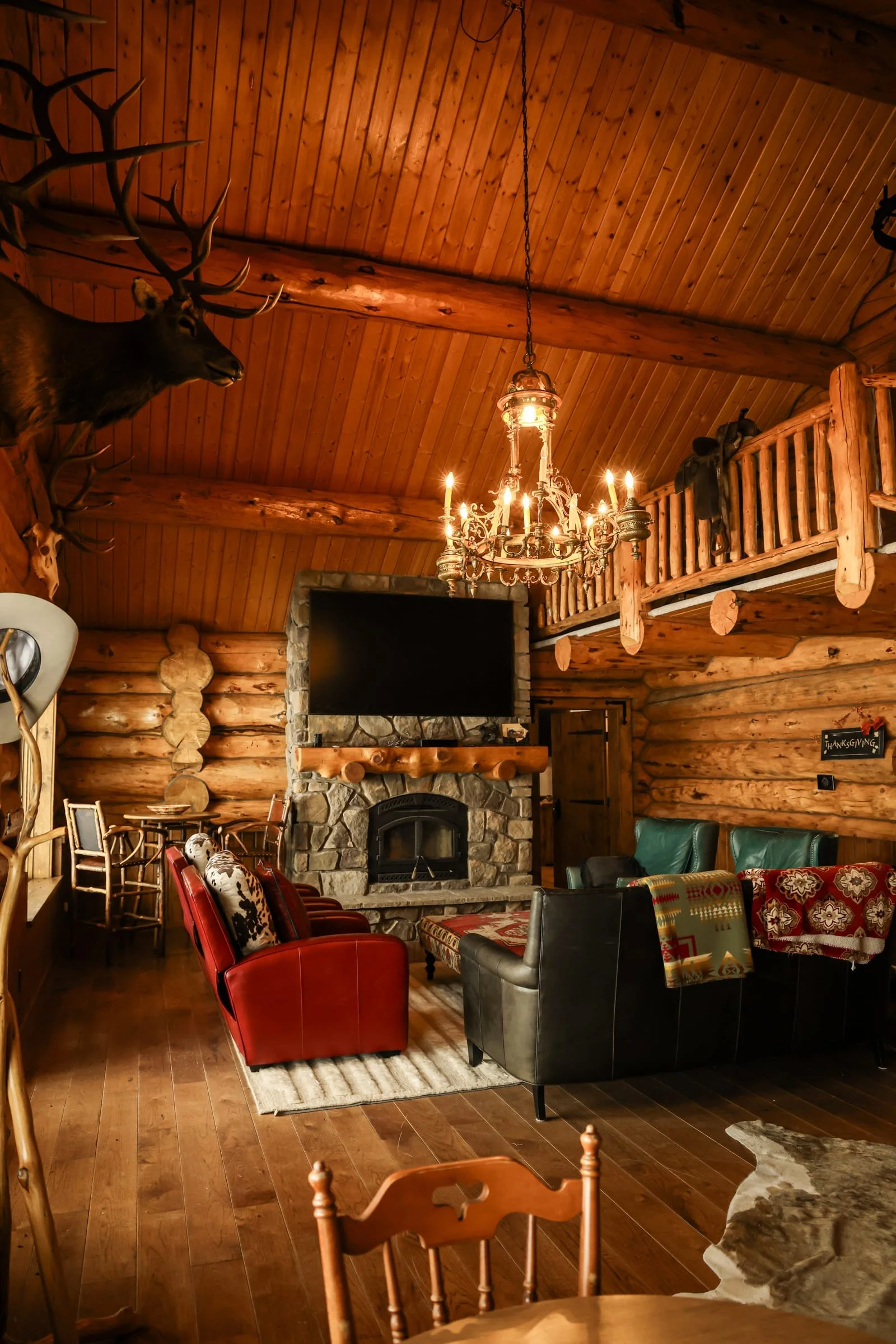 Interior of a rustic log cabin living room with wood-paneled ceiling, mounted deer and elk heads, stone fireplace, chandelier, and various chairs and sofas.