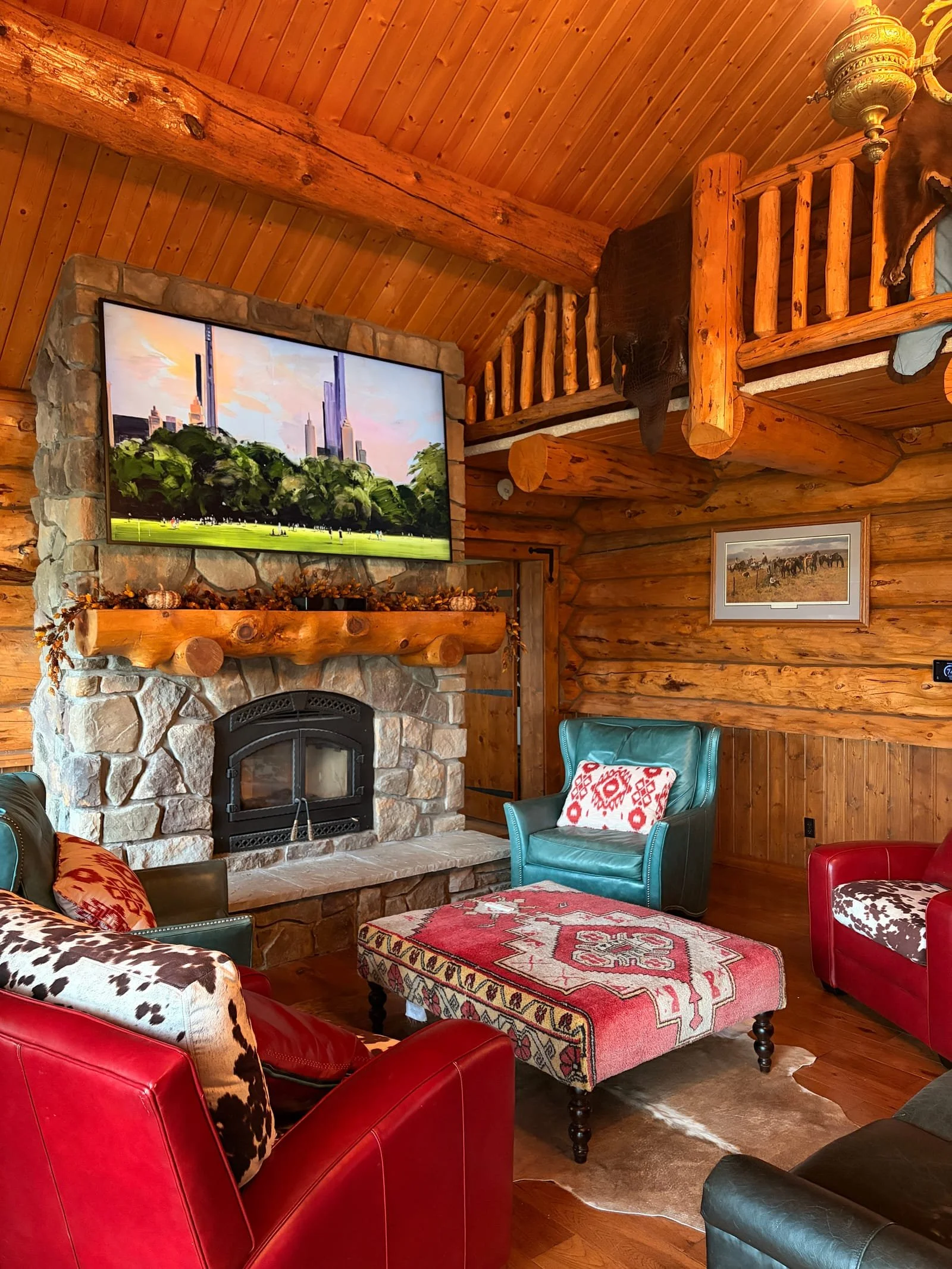 A cozy living room with wooden logs and paneling, a stone fireplace with a wooden mantle, multiple colorful chairs and a pink and white patterned ottoman, a wall art painting with trees and a city sky, and a portrait of elephants hanging on the wall.