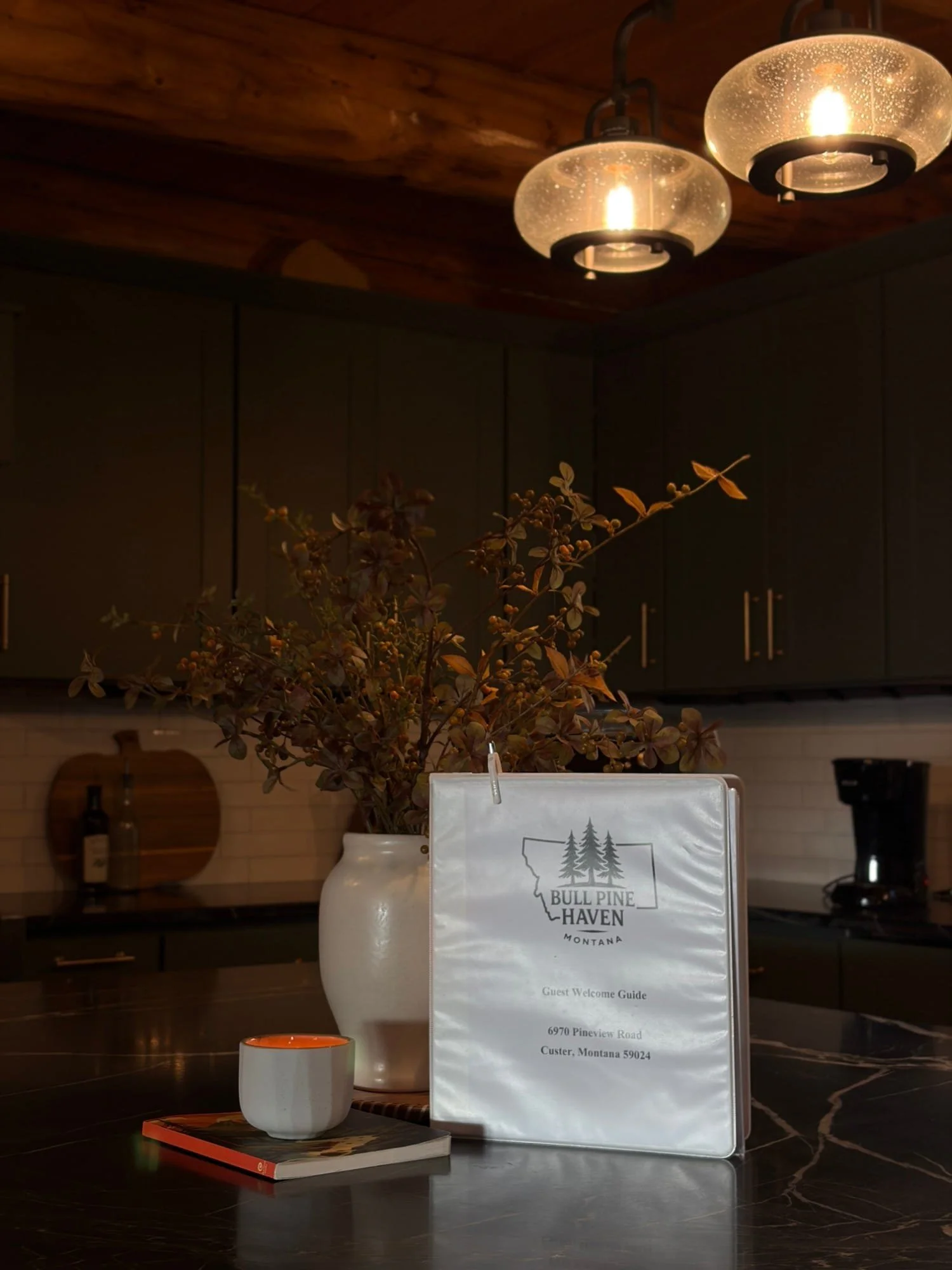 Interior of a rustic-themed room with wooden ceiling and modern lighting fixtures, featuring a large white vase with dried branches, a guest welcome guide with the logo 'Bull Pine Haven Montana,' a small orange mug on a book, and dark cabinets in the background.