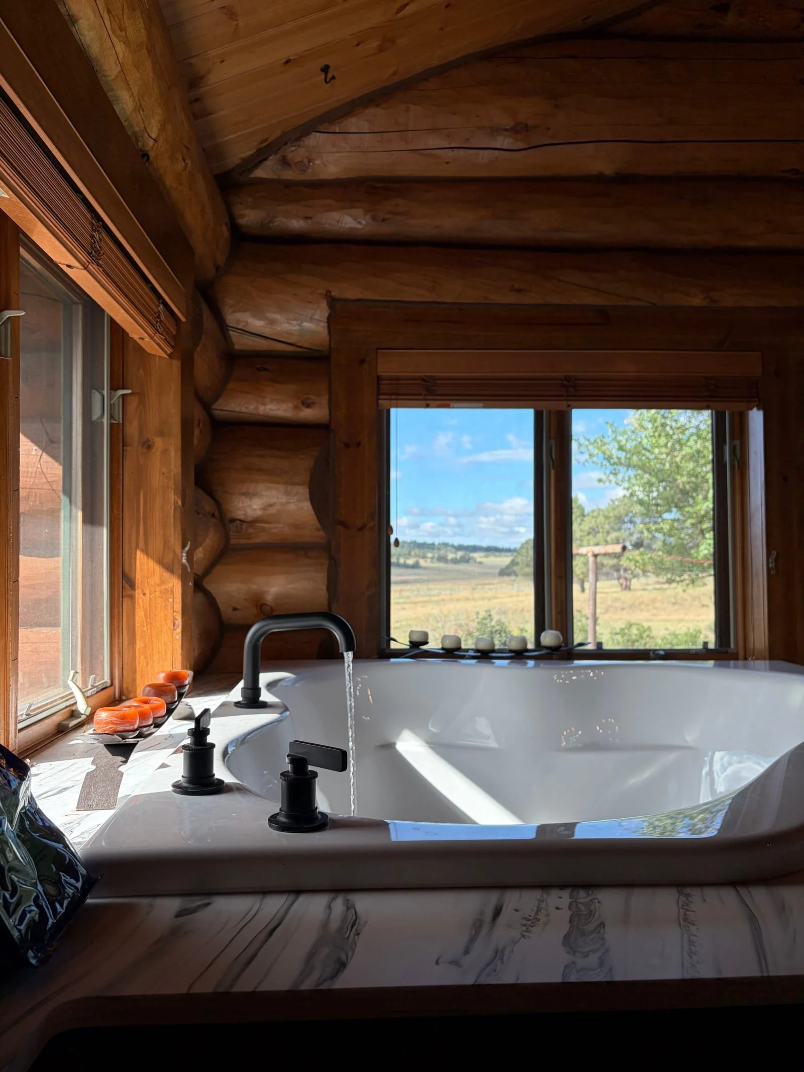 A bathroom with a large corner bathtub, black faucet and handles, a window showing an outdoor landscape with trees and open fields, and a wooden interior with log walls and a view of the countryside.