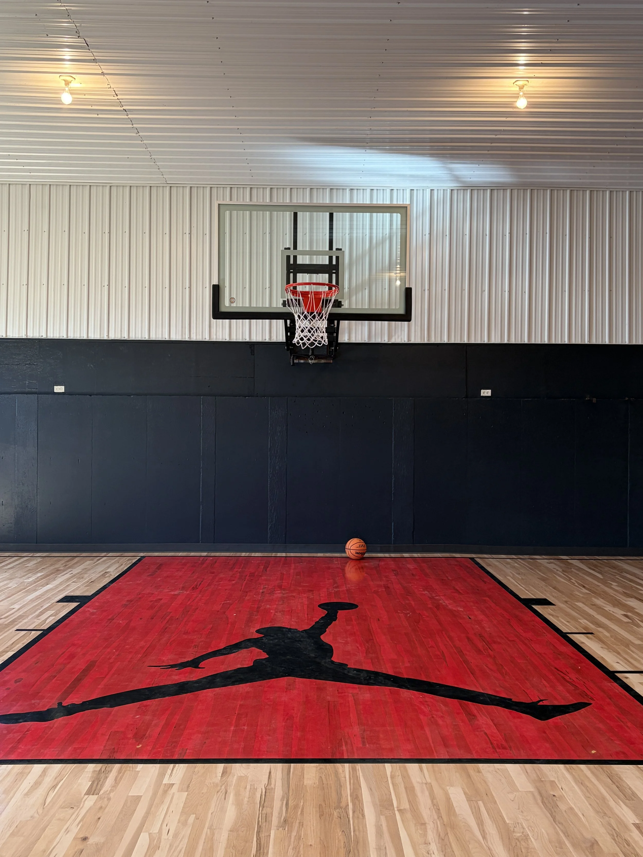 An indoor basketball court with a basketball hoop, a basketball, and a Red Jumpman logo painted on the wooden floor. The background features a wall with a glass backboard and unlit ceiling lights.