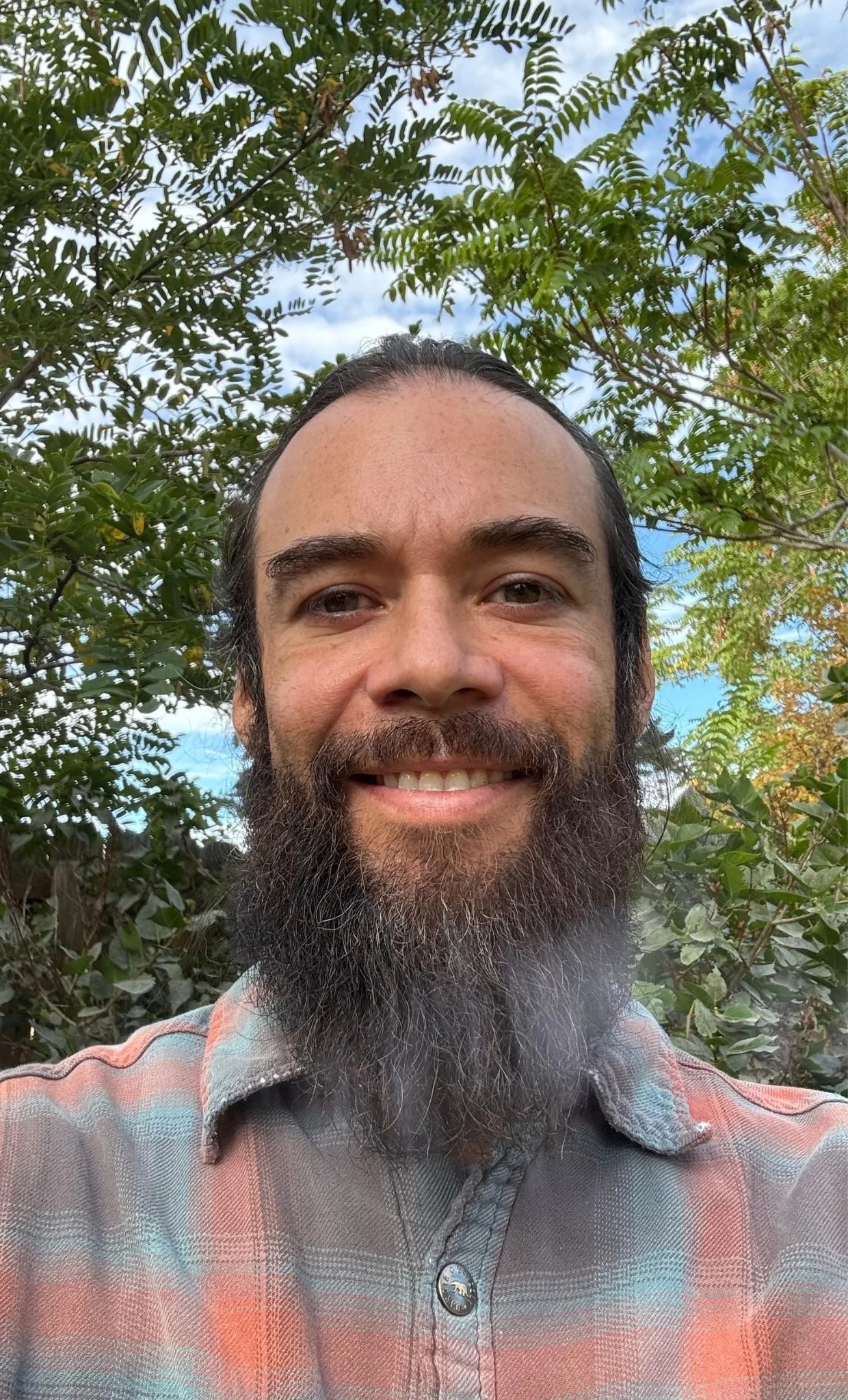 A man with long dark hair and a thick beard taking a selfie outdoors. Behind him, leafy green trees and a partly cloudy sky are visible.