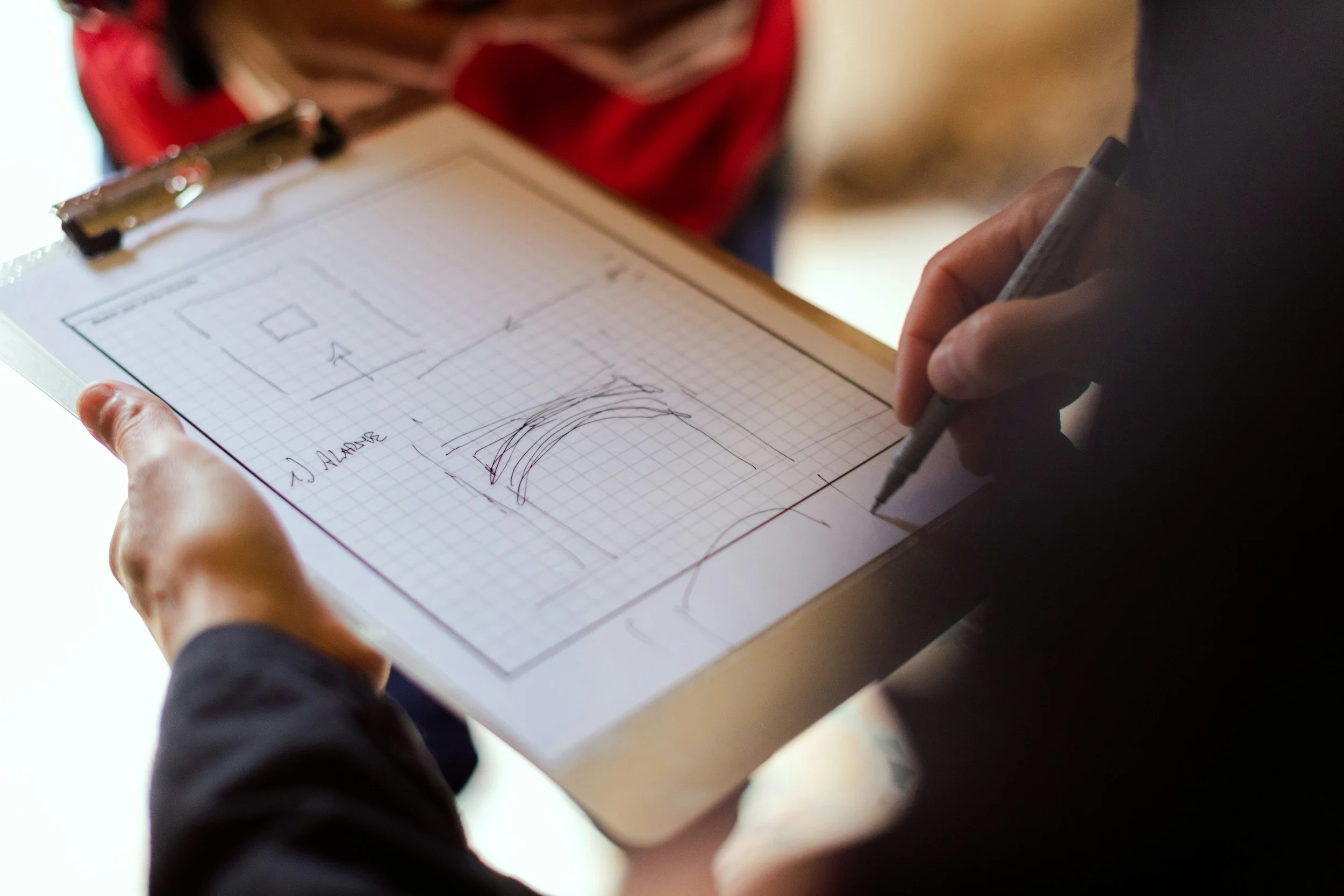 Person holding a clipboard with a grid paper and sketches of architectural designs, writing with a pen.