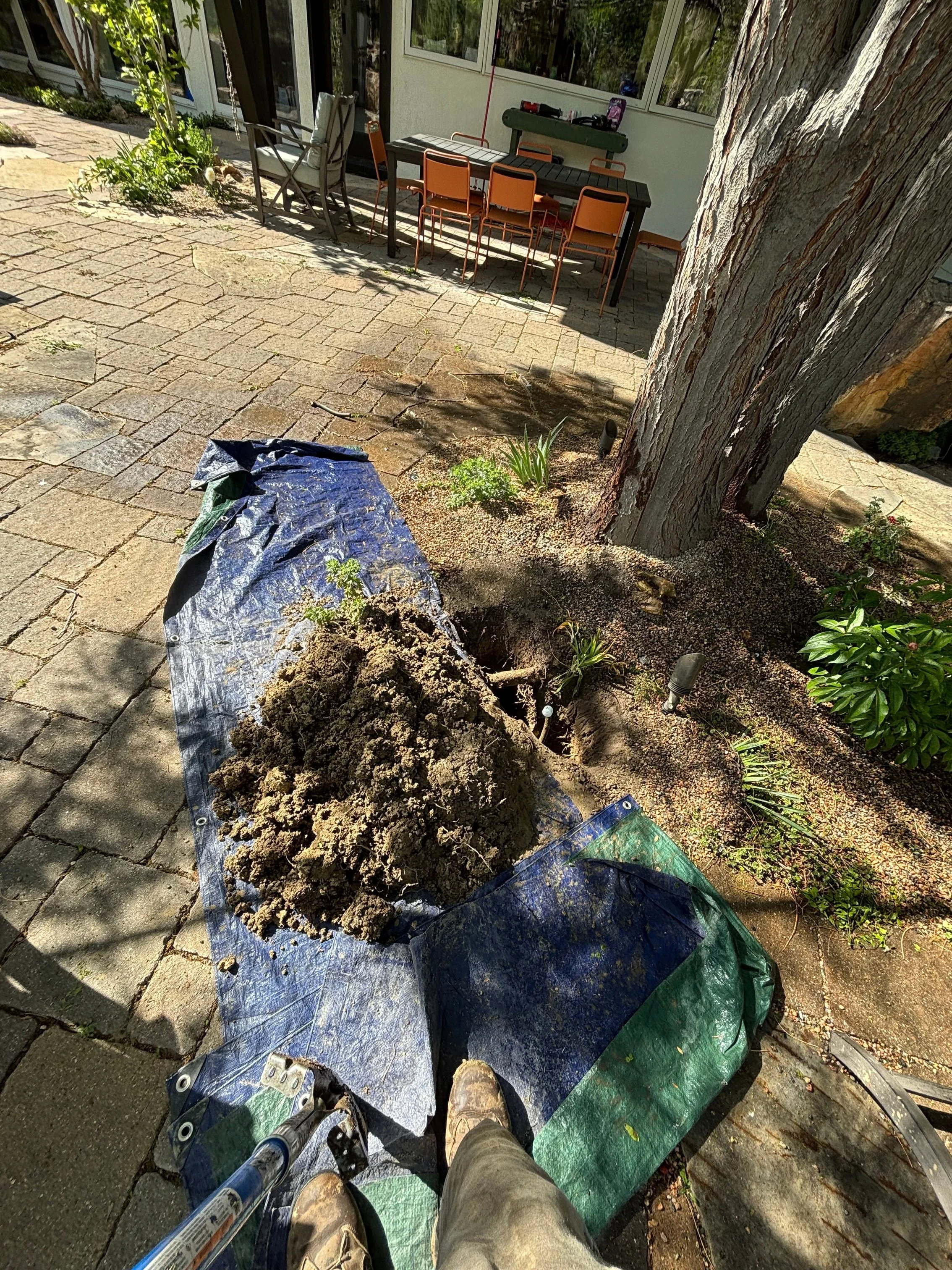 View from a person's perspective standing on a ladder or platform looking down at a garden work site with a pile of dirt, gardening tools, and a tarp on the ground, with outdoor furniture and trees in the background.