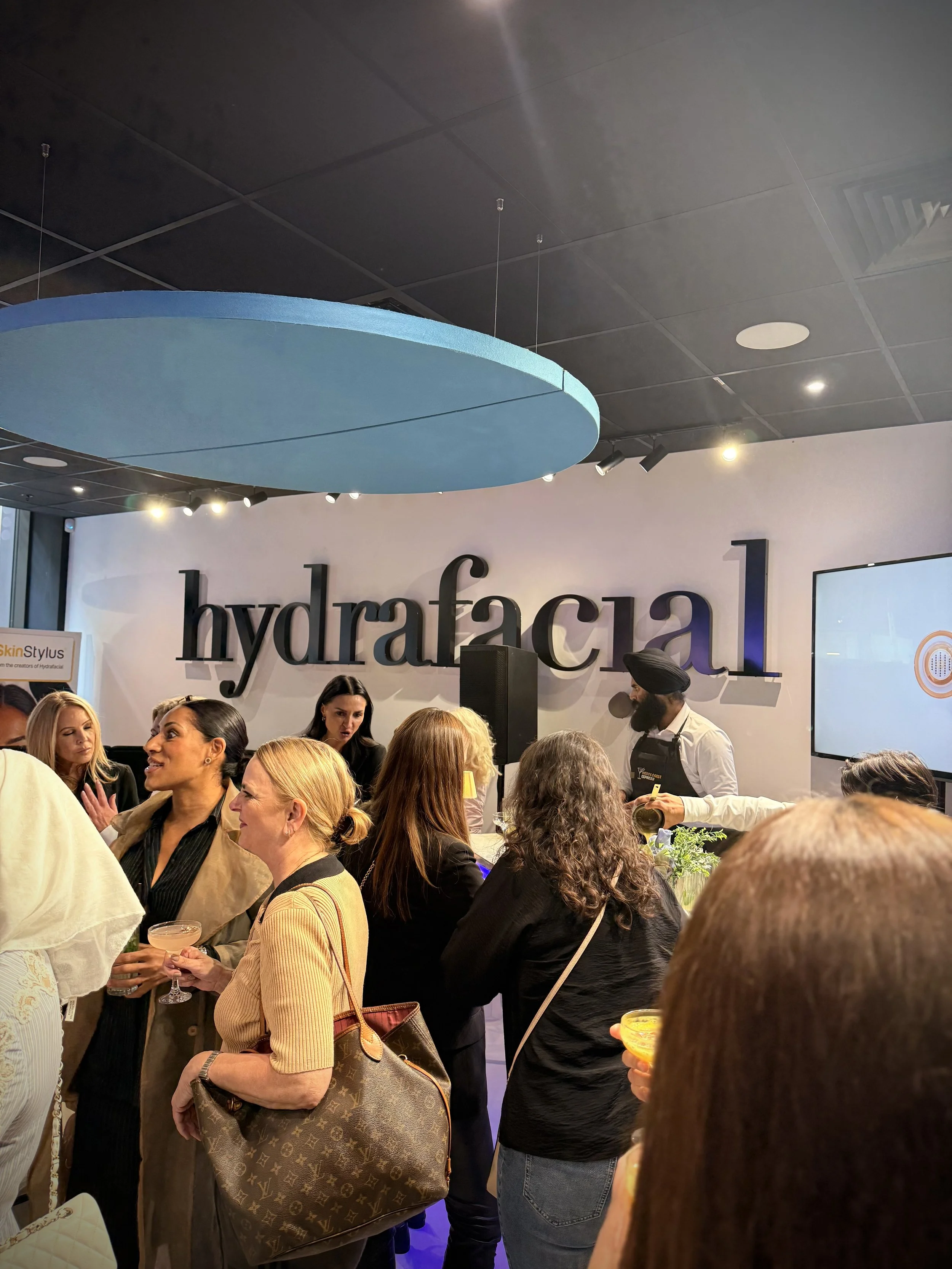 A group of women and a man gathered in a modern event space with a large sign that reads 'hydrafacial' on the wall. Some are holding drinks, and there is a bartender serving drinks in the background. The scene appears to be at a social or promotional event.