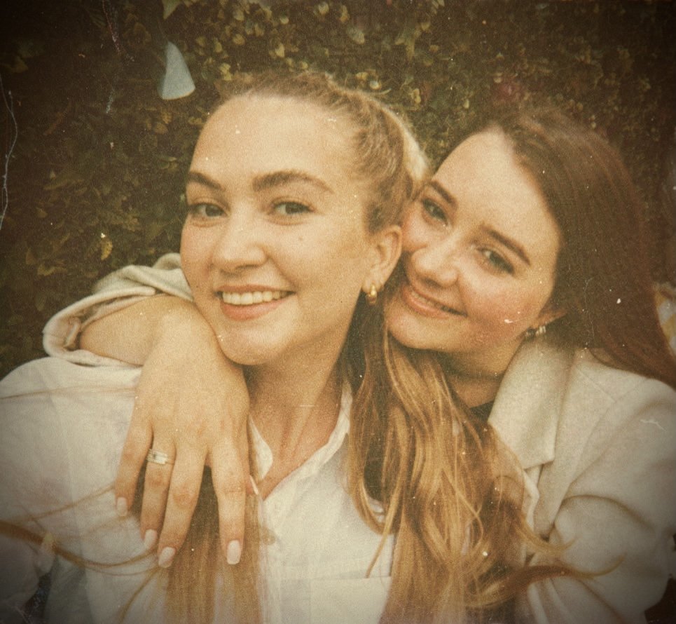 Two women smiling, one with blonde hair and the other with brunette hair, hugging outdoors amidst foliage.