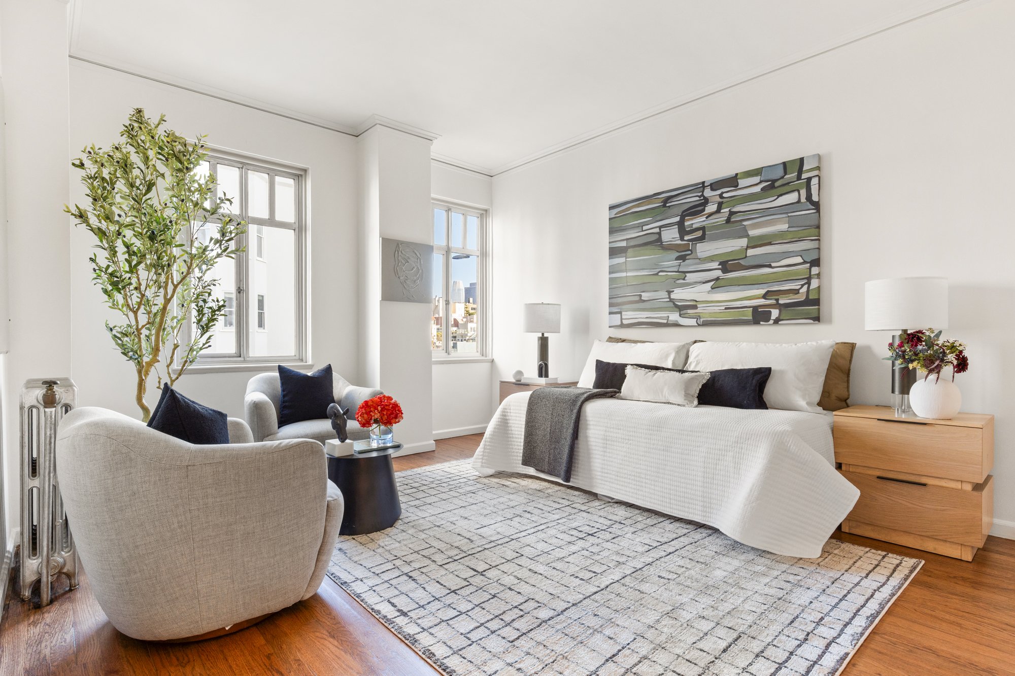 Bright modern bedroom with white walls and large windows, featuring a beige armchair with navy pillows, a bed with white bedding and dark pillows, a wooden nightstand, and a large abstract painting on the wall.