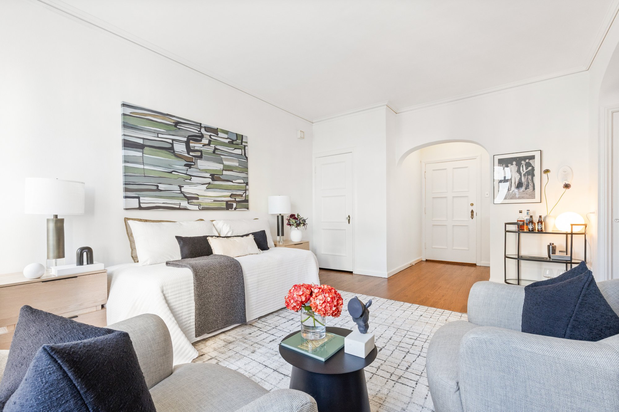 Modern living room with white walls, a white sofa, dark gray armchairs, and abstract artwork on the wall. The room features a small black side table with decorative items and a vase of flowers, hardwood floors, and some framed photos or art.