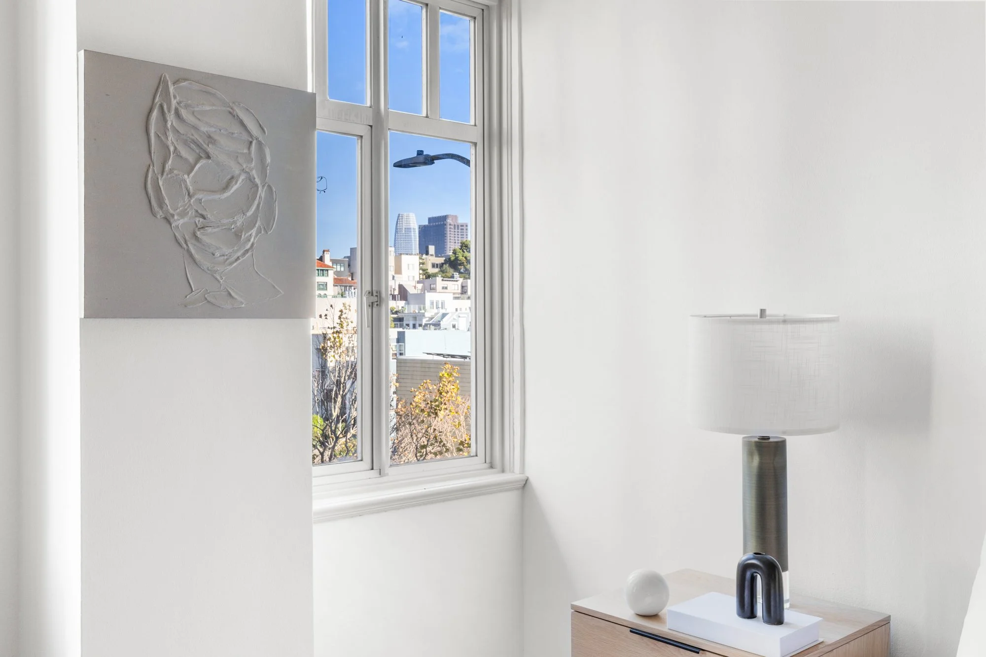 A minimalist indoor scene featuring a white wall with a modern abstract artwork and a large window showing a cityscape with buildings and a blue sky. A floor lamp and a wooden furniture piece with decorative objects are also visible.