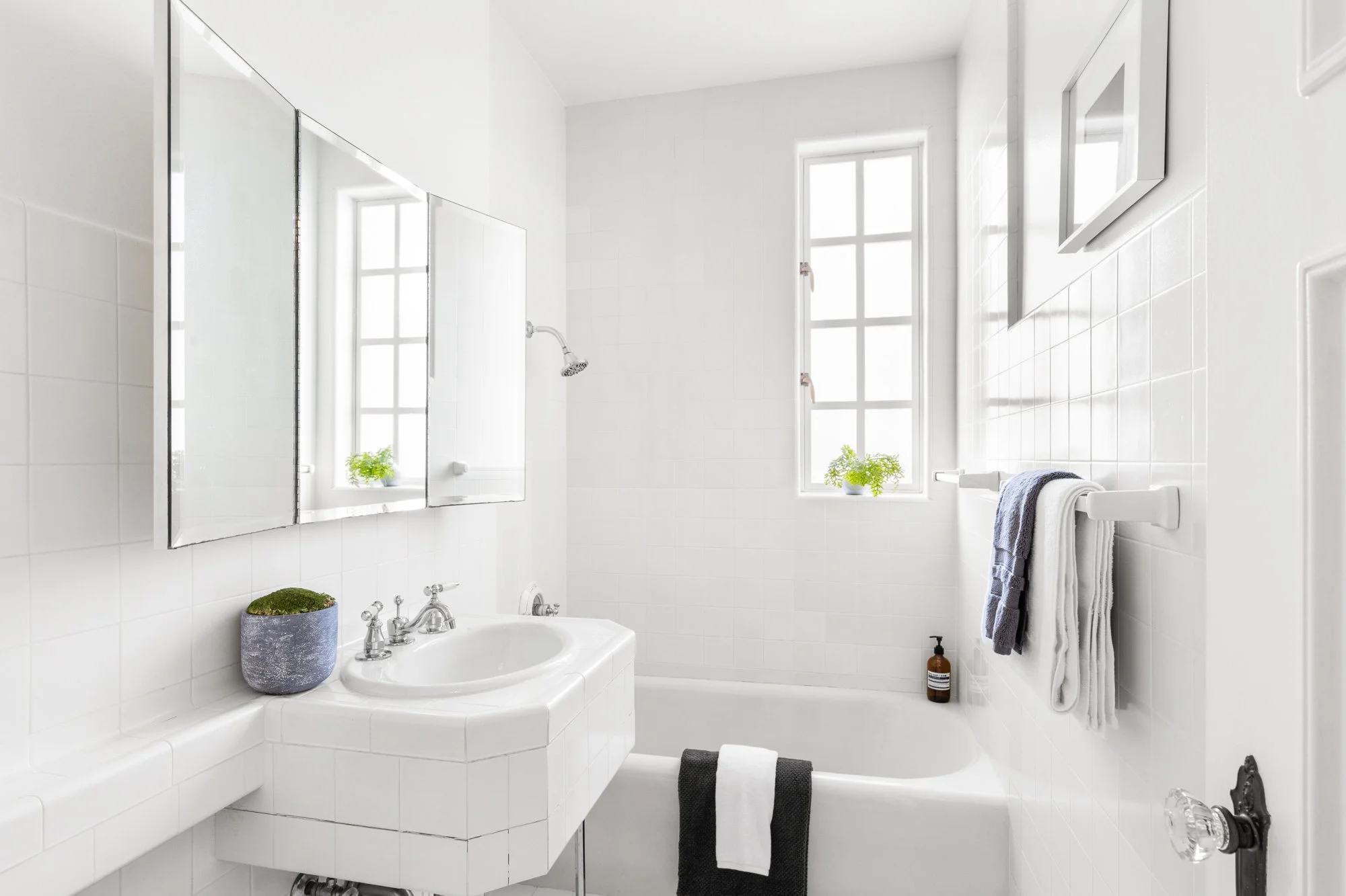 Bright bathroom with white tiled walls, a bathtub with a black towel, a white towel rack with towels, a window with plants, a mirror, a small plant on the sink, and a bottle of soap.