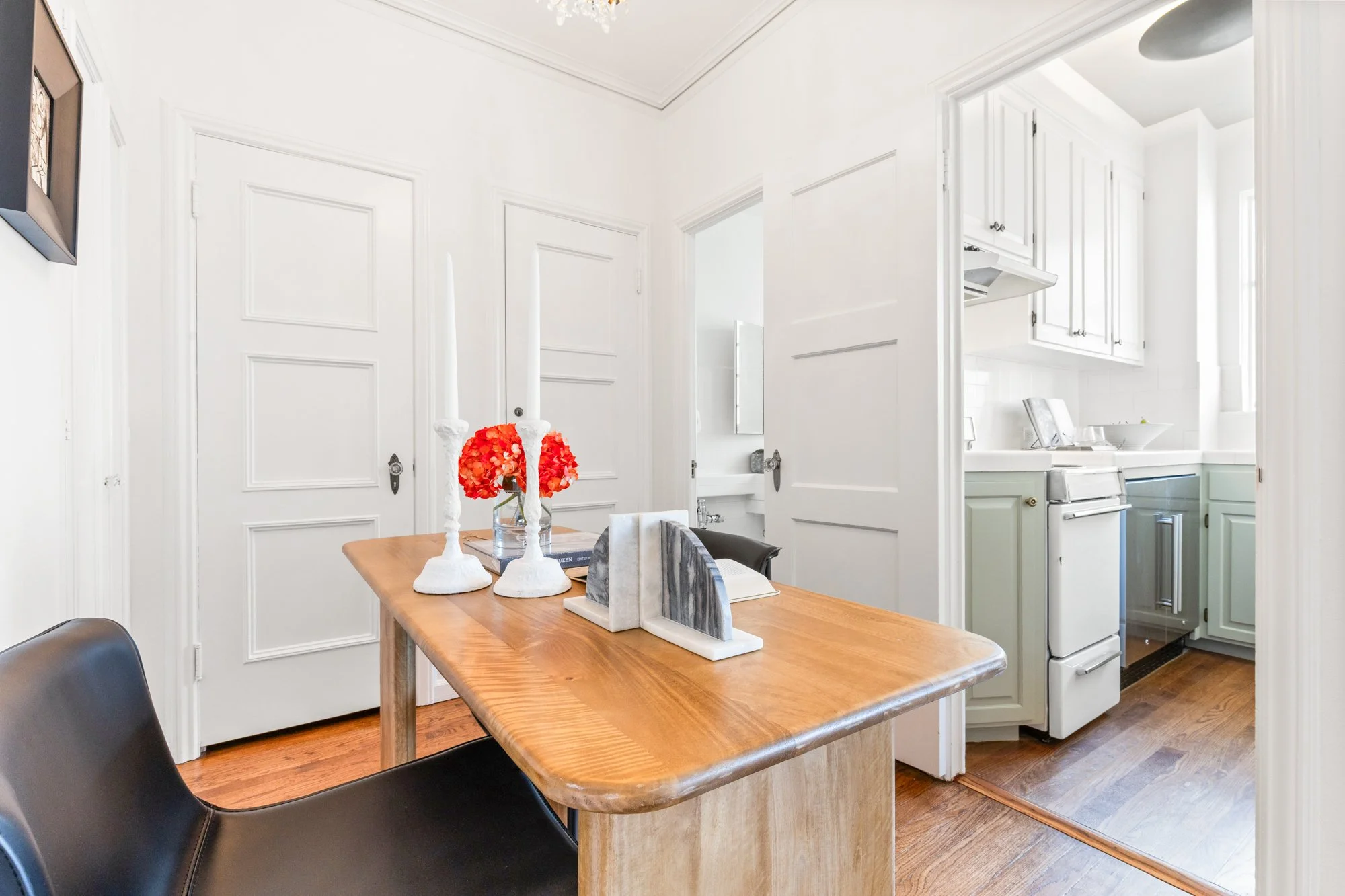 Bright dining area with a wooden table and black chairs, decorated with white candle holders and red flowers, adjoining a white kitchen with cabinets and appliances.