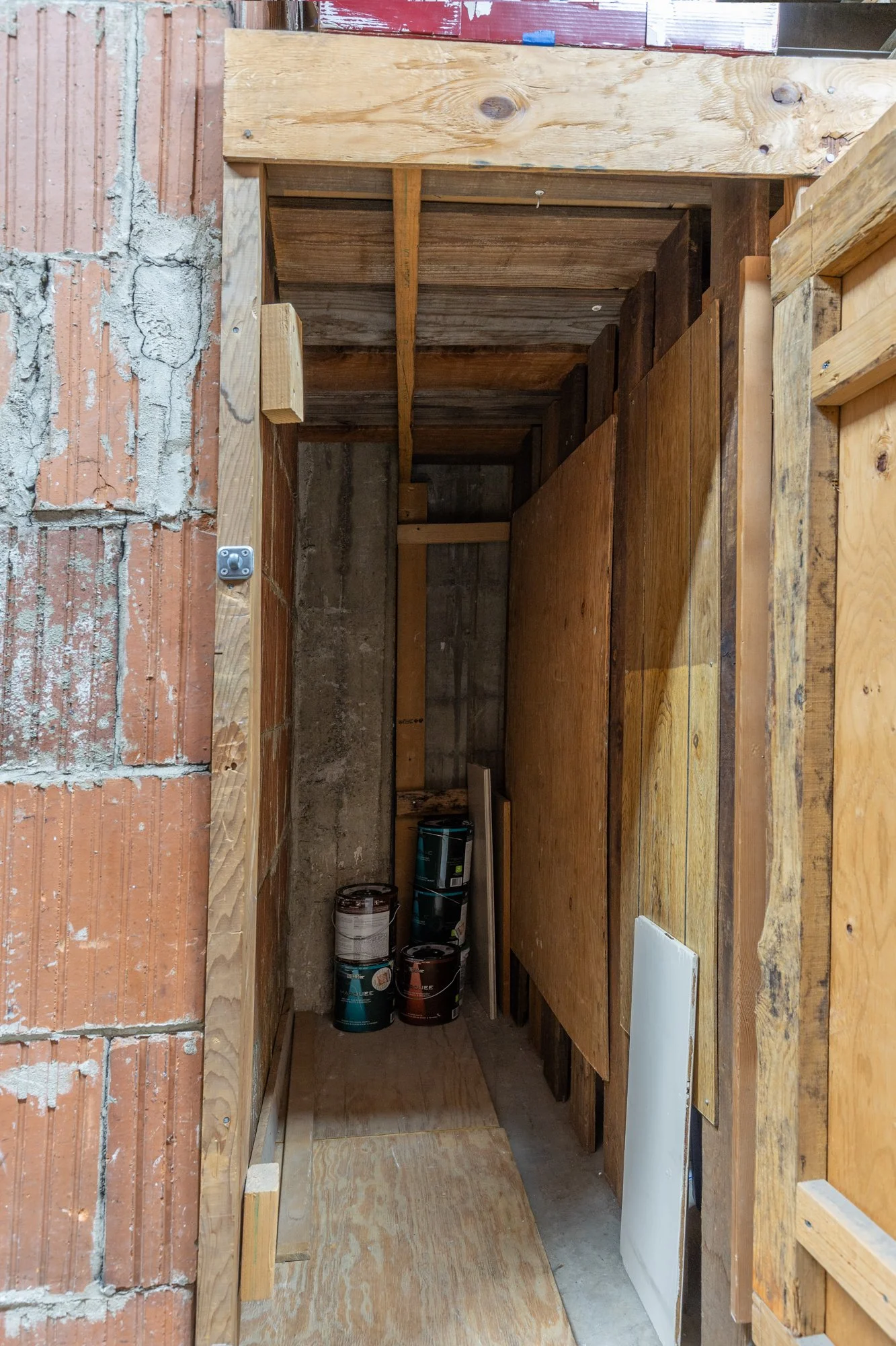 Construction site with wooden framing, ladders, and paint cans stored in a narrow space between brick and wooden walls.