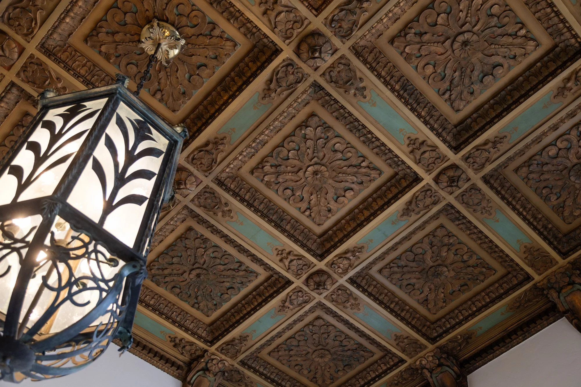 Decorative wooden ceiling with intricate floral and geometric carvings, illuminated by a black metal lantern with a stained glass panel and ornate design.