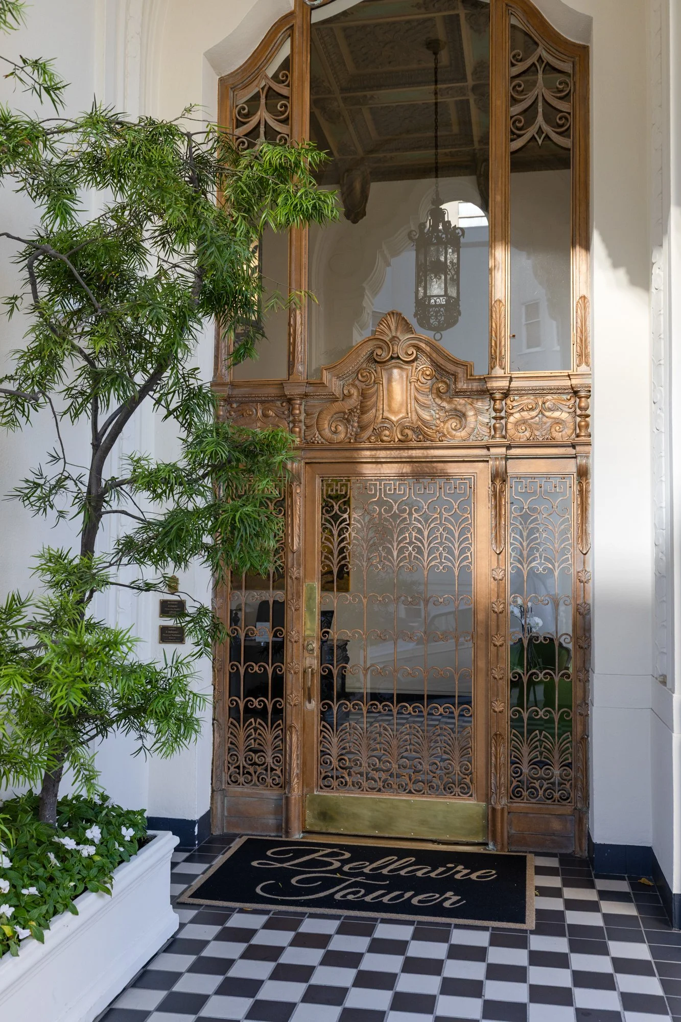 Elegant elevator door made of ornate woodwork with a glass window, inside a building with decorative ceiling and a chandelier, next to a green plant and flowers, with a welcome mat on a black and white checkered floor reading 'Bellvue Tower.'