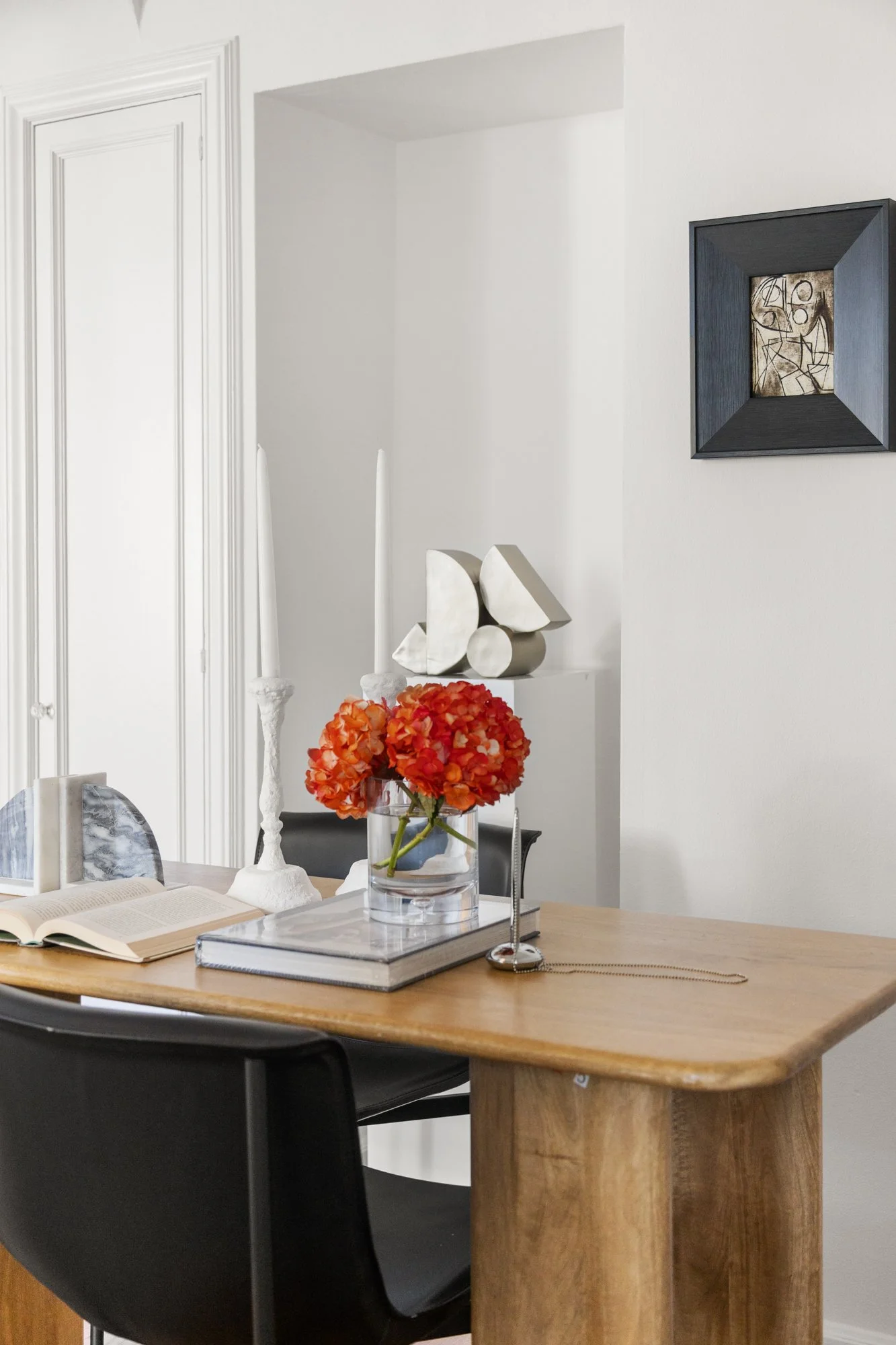 A wooden dining table with a glass vase of orange flowers, an open book, and a tray with a decorative object. Part of a black chair and a white decorative sculpture are visible. The background features a white wall with a framed abstract artwork and 