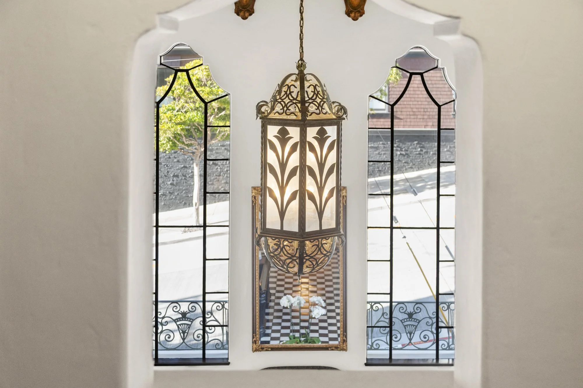 Ornate hanging lantern with intricate metalwork, suspended in front of arched windows with black metal frames, overlooking a courtyard with checkered flooring and potted white orchids.