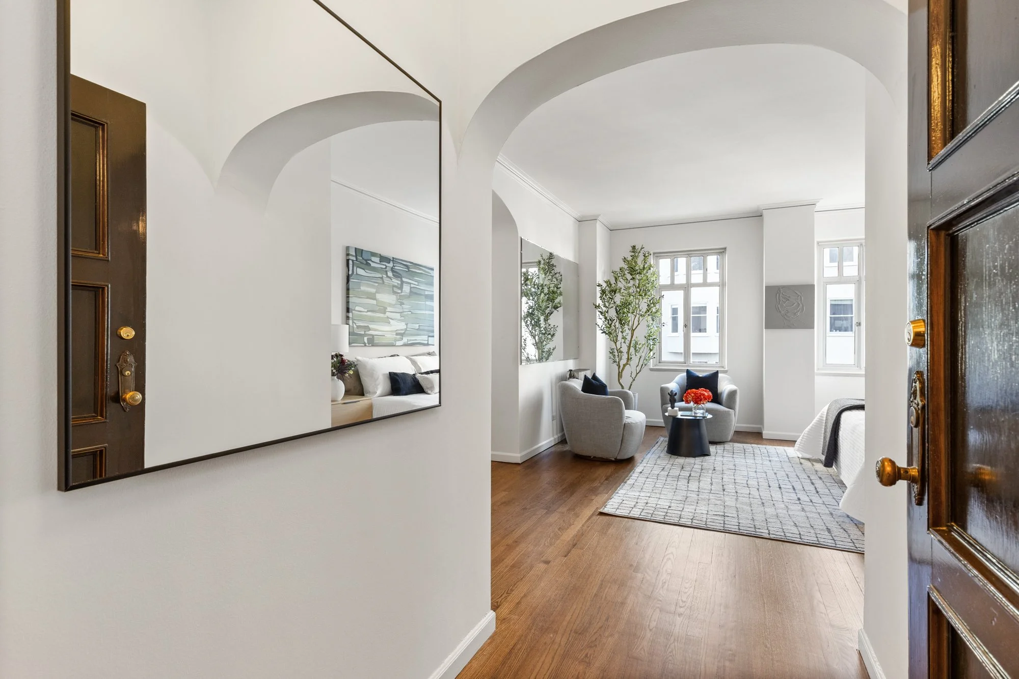 Living room with two light-colored armchairs, a small round black side table with a vase of red flowers, large windows, potted plants, abstract wall art, and a patterned area rug.