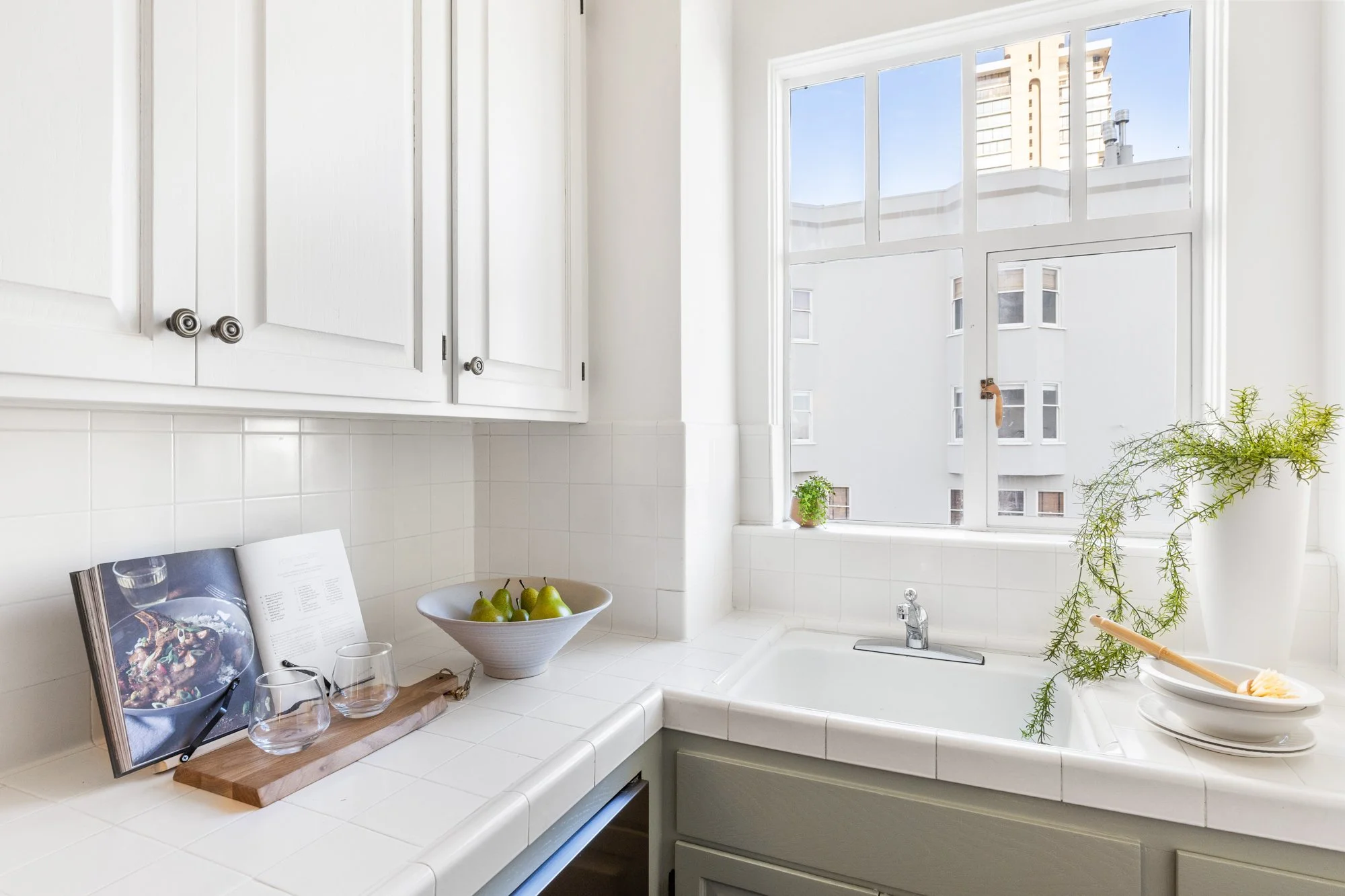 Kitchen with white cabinets, a large window with a city view, a white sink, and a countertop featuring a bowl of green pears, a cookbook, wine glasses, and some potted plants.