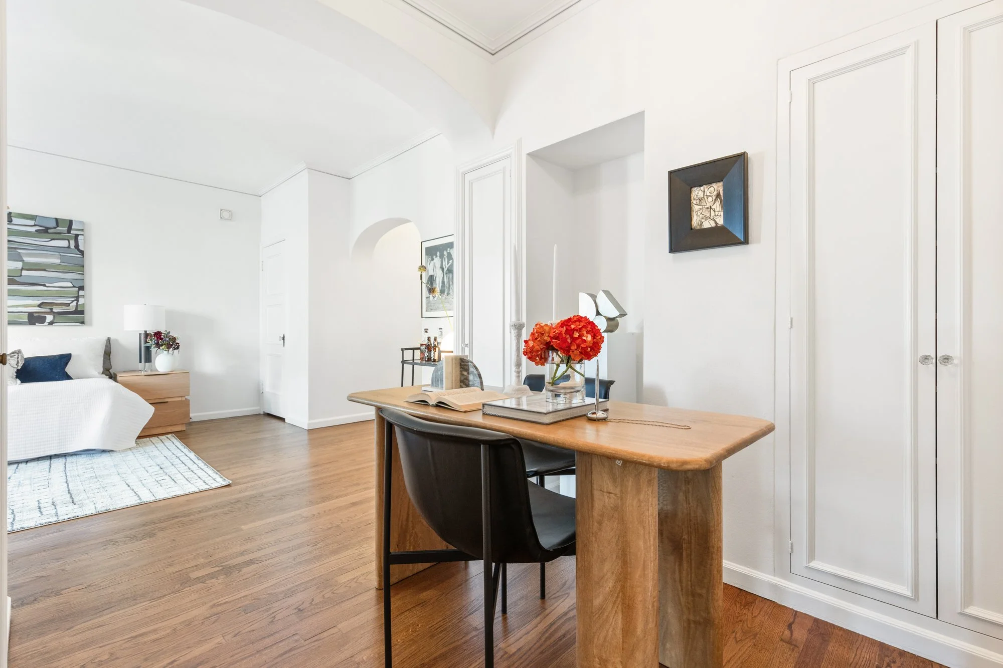 A bright living space with white walls and wooden flooring, featuring a bed with a colorful painting hung above it, a nightstand with a white lamp and flowers, and a dining table with a black chair, decorative objects, and a vase of red flowers.