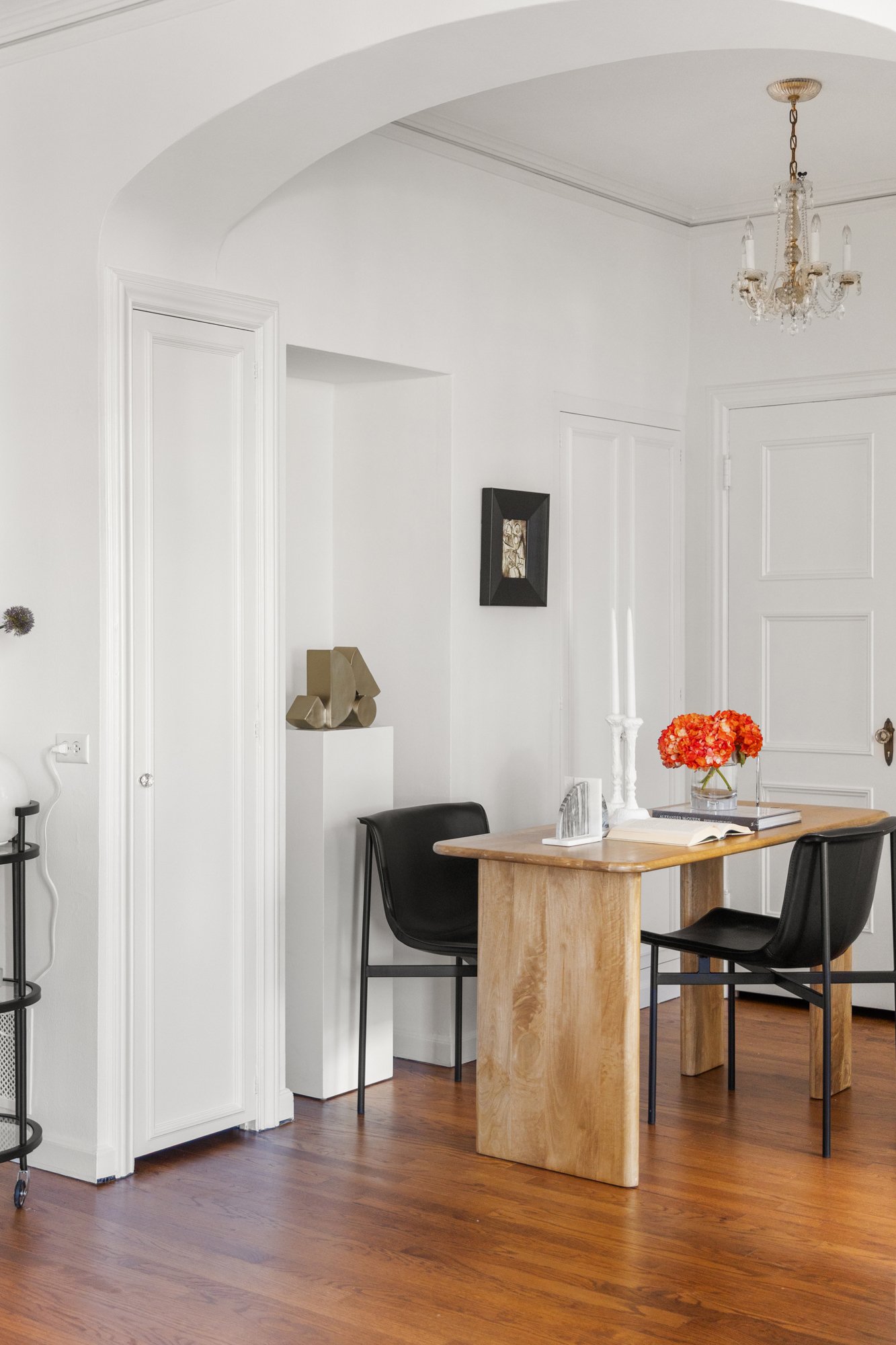 Interior of a room with white walls, a wooden dining table, black chairs, a floral centerpiece, a chandelier, and a small framed art piece.