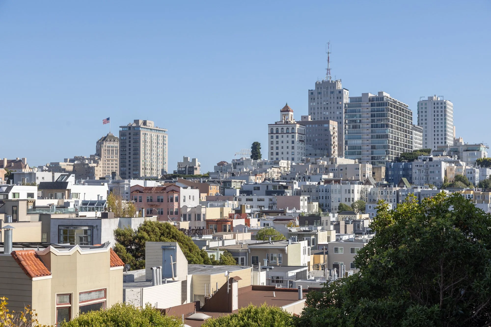 A city skyline with a mix of modern and historic buildings under a clear blue sky. Residential houses are visible in the foreground, with taller buildings and skyscrapers in the background. A flag is flying on one of the buildings.