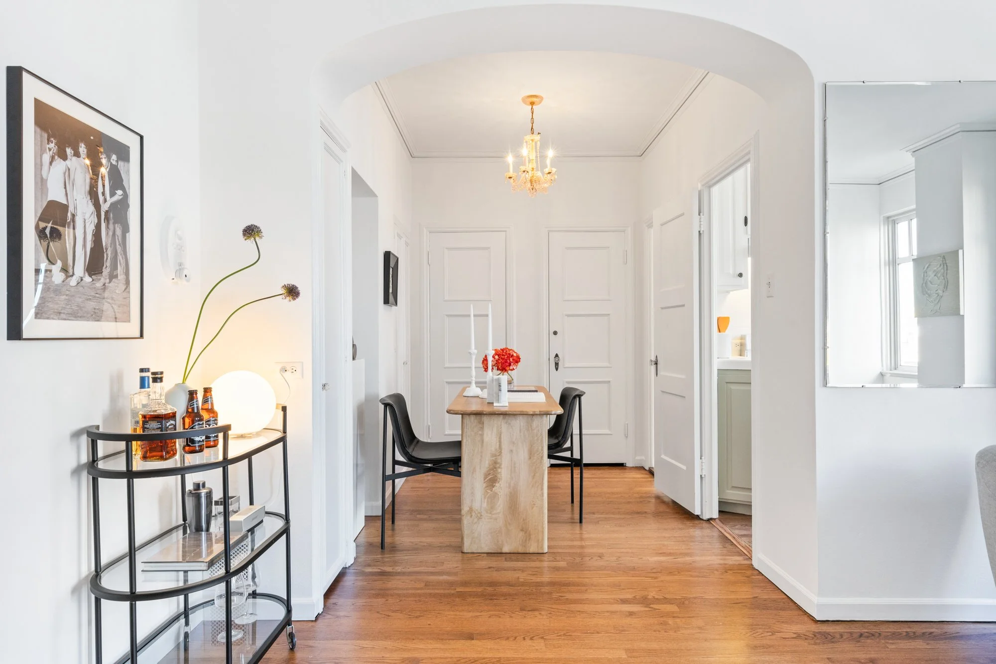 Bright, white-walled dining area with a small wooden table, black chairs, and decorative lighting, including a chandelier and wall-mounted lights, with artwork on the walls and a bar cart holding bottles of alcohol.
