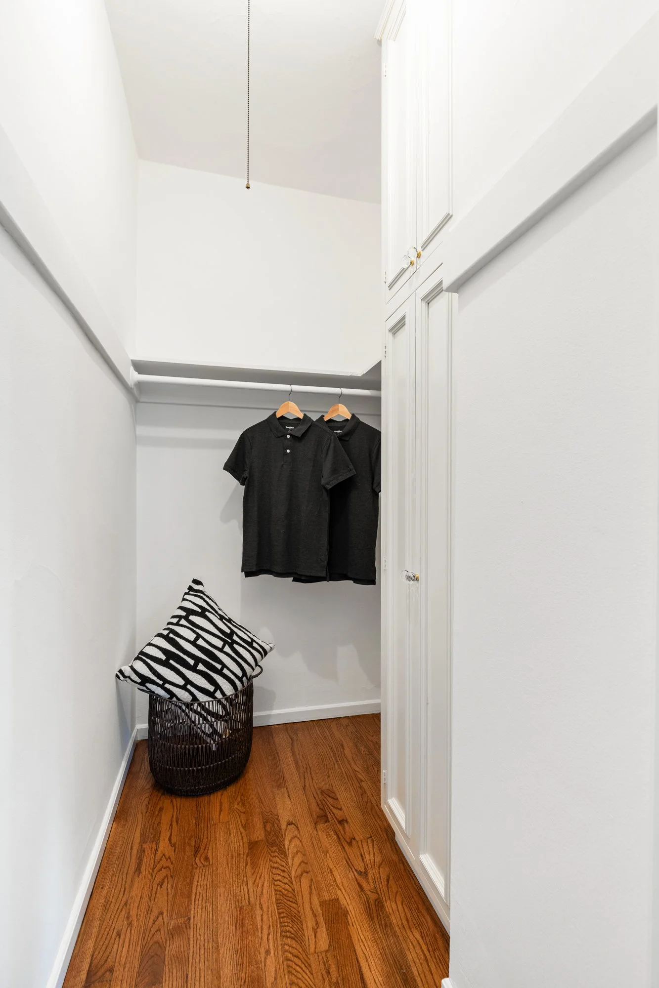 A small walk-in closet with white walls and wood flooring. Two black polo shirts hang on hangers suspended from a white rod. There is a black wicker basket with a black and white patterned cushion on top, positioned at the corner of the closet.