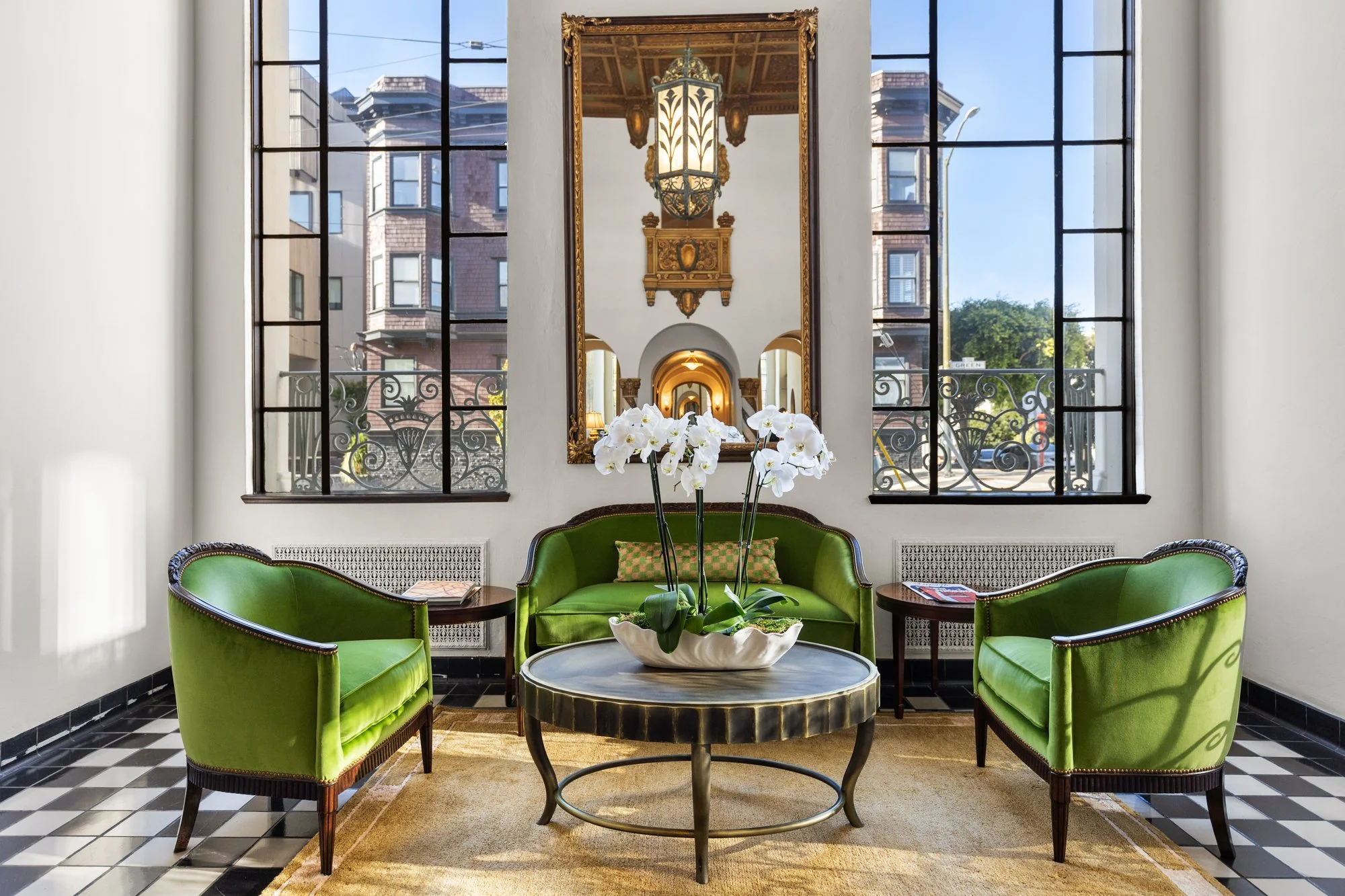 Interior of a room with green velvet armchairs surrounding a round coffee table with a white orchid plant. Large windows with black framing let in natural light, and a large mirror reflects a hanging lantern and ornate architectural details. A window