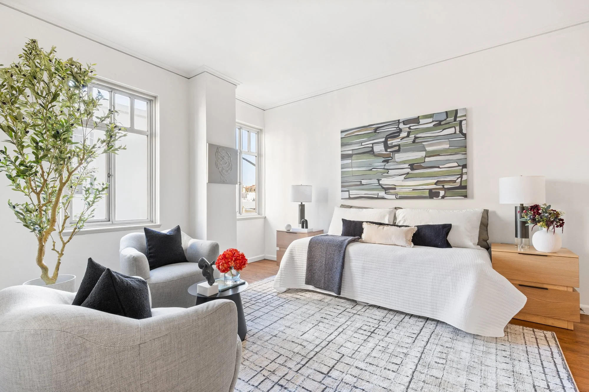 Living room with white walls, large windows, a cream sofa with black and beige pillows, a white armchair with black pillows, two side tables with lamps, an abstract painting on the wall, a potted plant, and a patterned rug.