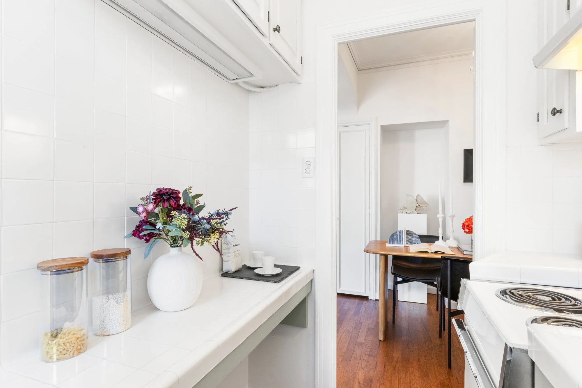 A white kitchen with a white tiled wall, a countertop with a large white vase holding a colorful bouquet of flowers, glass containers with pasta, and a tray with cups. In the background, there is a small wooden table with an open book, candles, and f