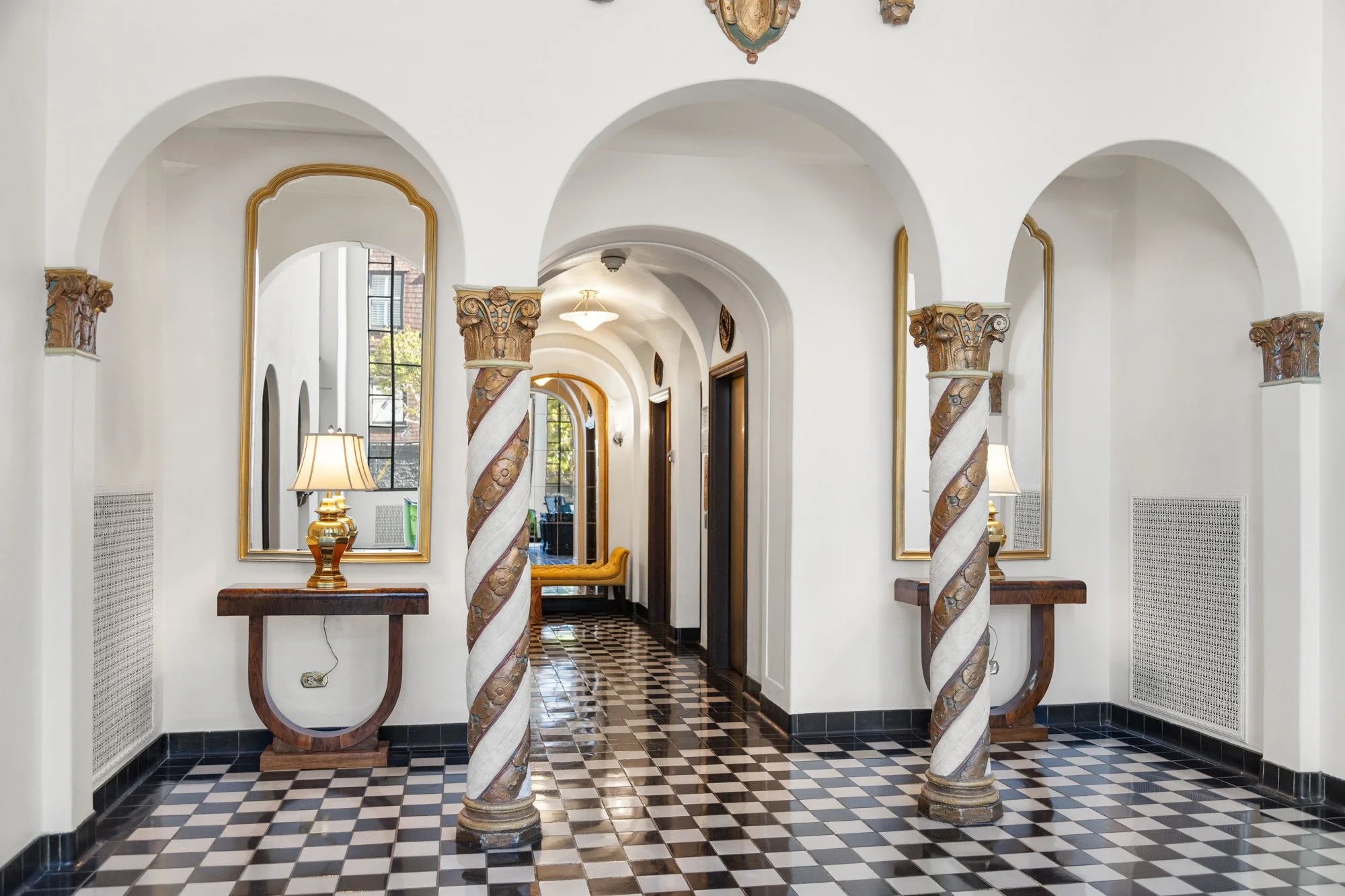 Interior view of a hallway with arched doorways, decorative columns, black and white checkered floor, and mirrors reflecting lamps and furniture.