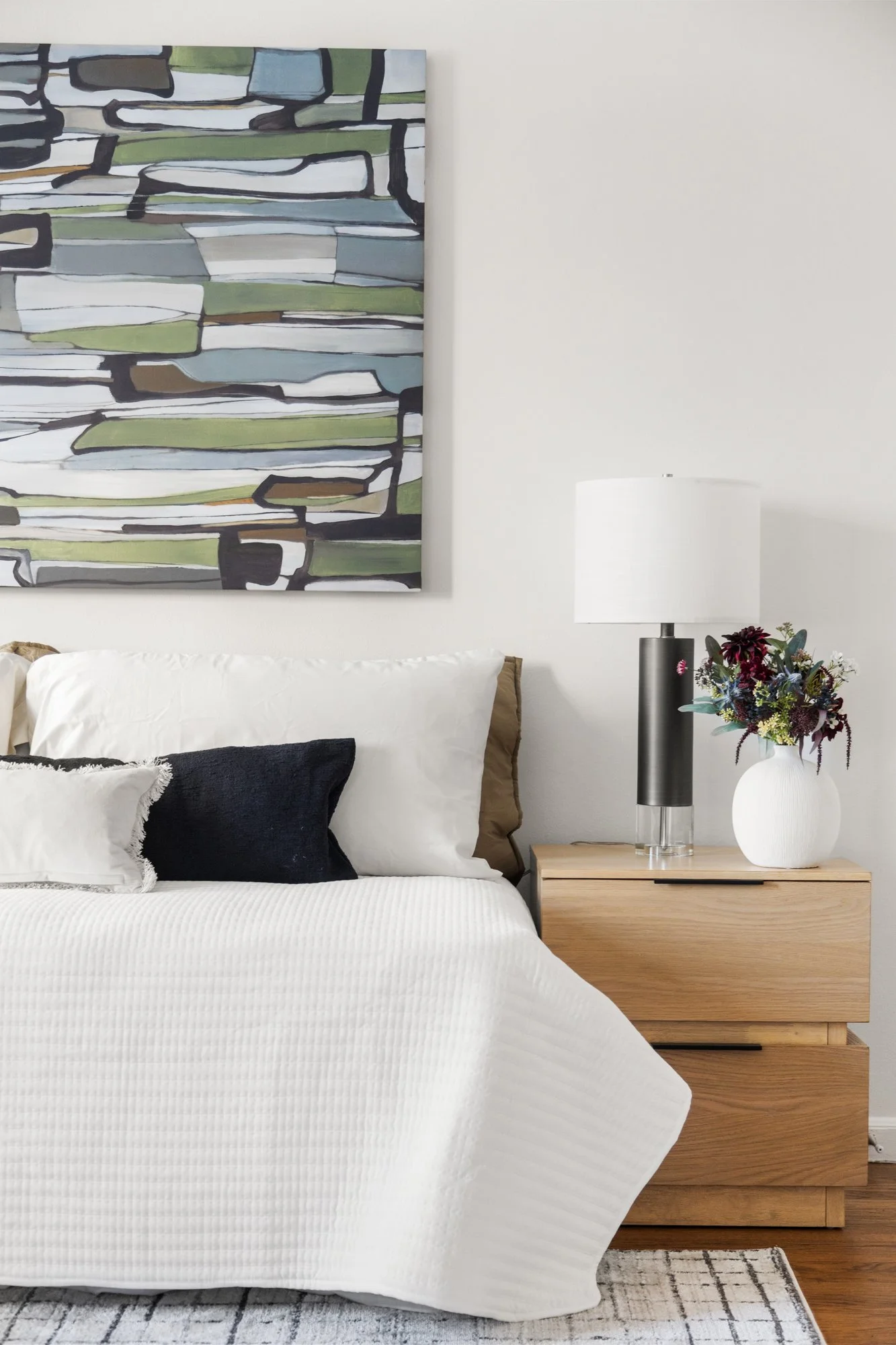 A modern bedroom with a white bed, black and white pillows, a wooden nightstand with a lamp and a vase of flowers, a large abstract painting on the wall, and a rug on hardwood floor.