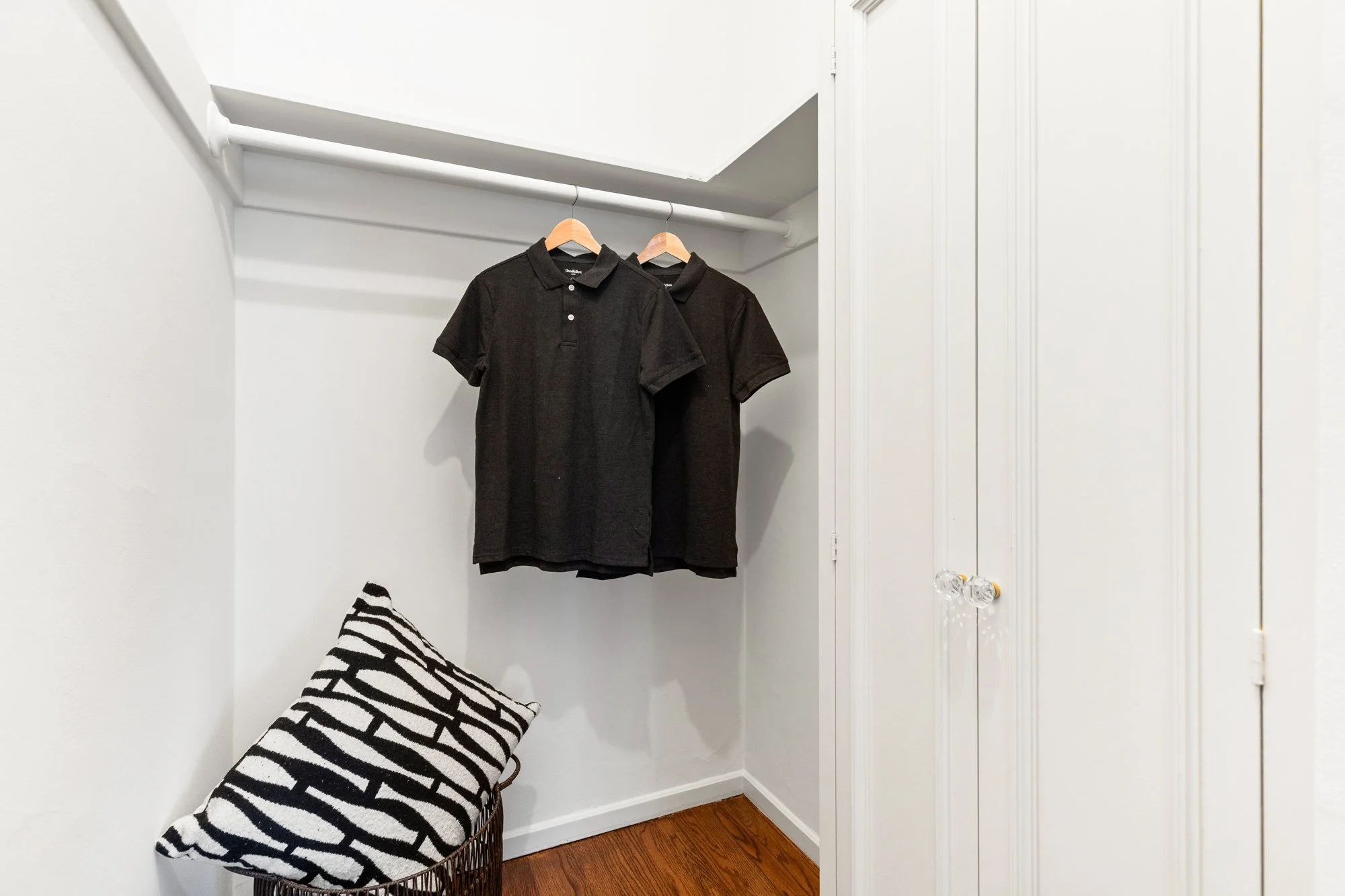 A small walk-in closet with two black polo shirts hanging on wooden hangers on a rod. There is a corner bench with a black and white striped pillow, and a white closet door with two knobs. The floor is wooden.