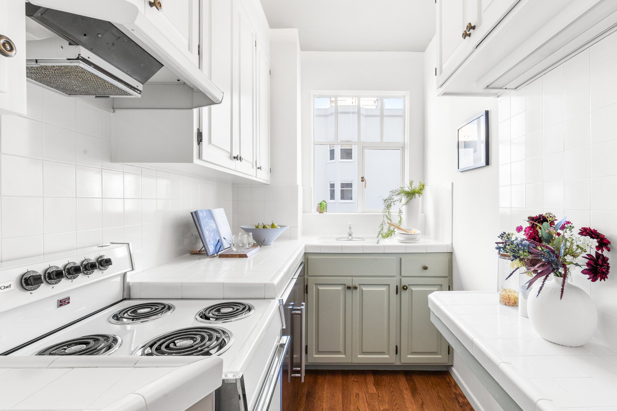 A compact, white kitchen with white cabinets, a white tile countertop, and a white stove with four burners. There is a window above the sink with sunlight streaming in, and a small plant and flowers decorate the right side of the counter.