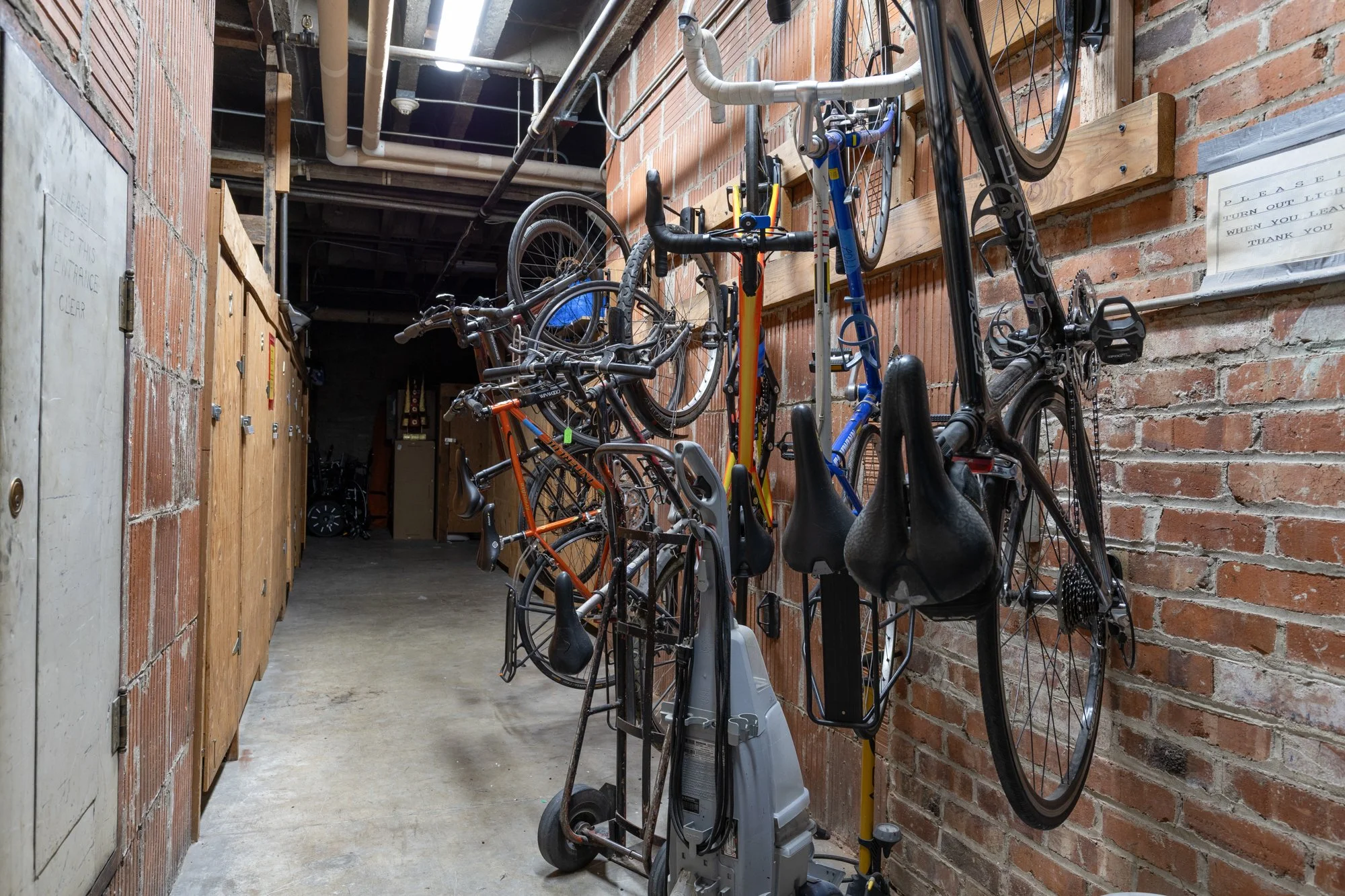 Indoor bike storage area with bikes mounted on a brick wall, a scooter, and equipment in a narrow hallway with concrete floor and wooden lockers.
