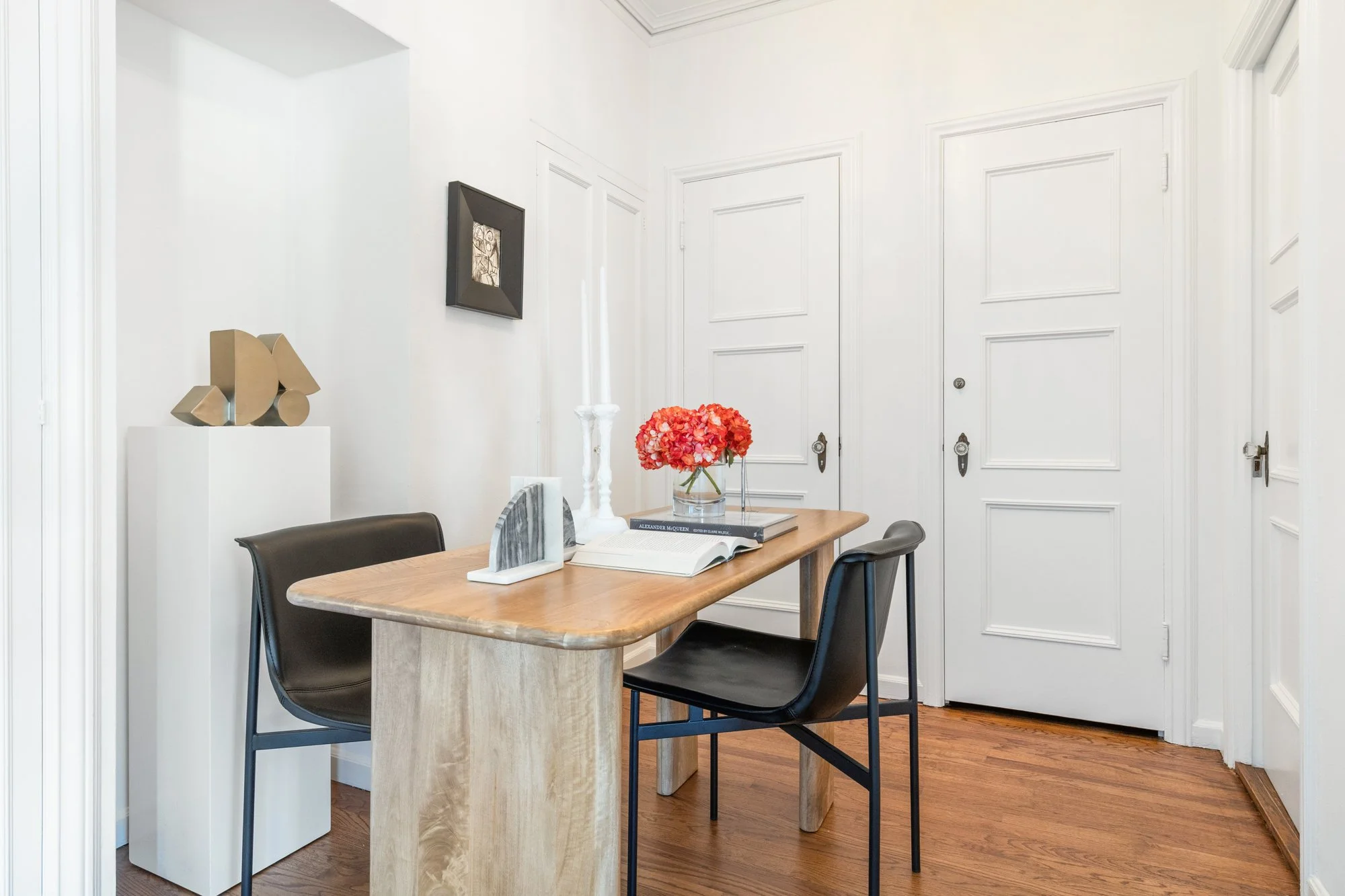 Interior of a room with white walls, a wooden table, with a red flower bouquet, books, and decorative items on the table, black chairs, white doors, artwork on the wall, and a wooden floor.