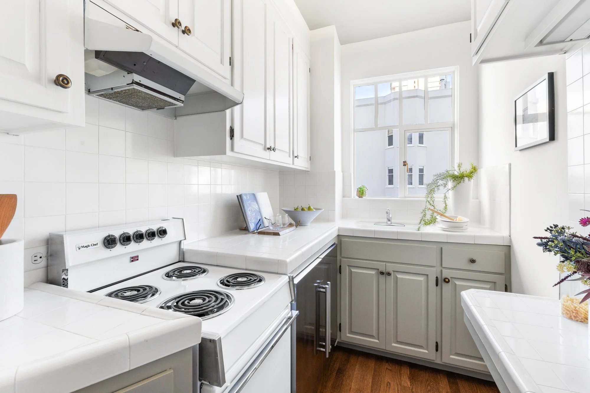 A small, white kitchen with white cabinets, a white countertop, white tile backsplash, and a window with a view of a neighboring building. The stove has four coil burners, and there are various kitchen items including a bowl of pears, a cookbook, gla