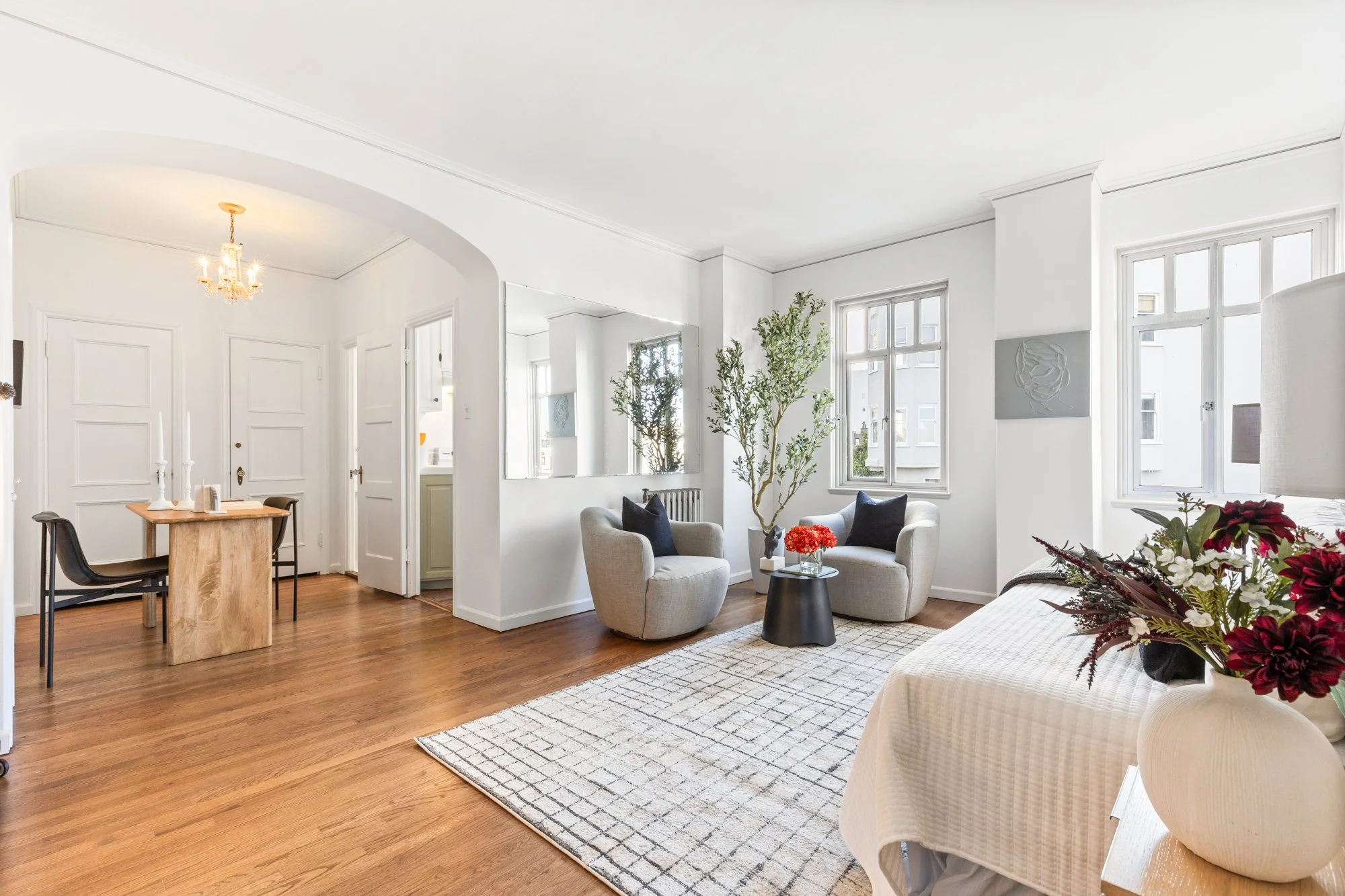 Bright living room with white walls, large windows, hardwood floors, and modern furniture including a sofa, armchairs, and a coffee table with flowers. A dining area with a wooden table and black chairs is visible.